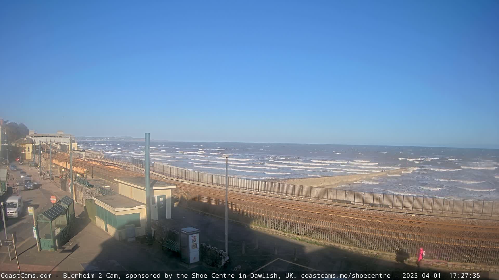 A coastal scene under a clear blue sky shows a train track running alongside a beach with moderate waves and a sea wall.
