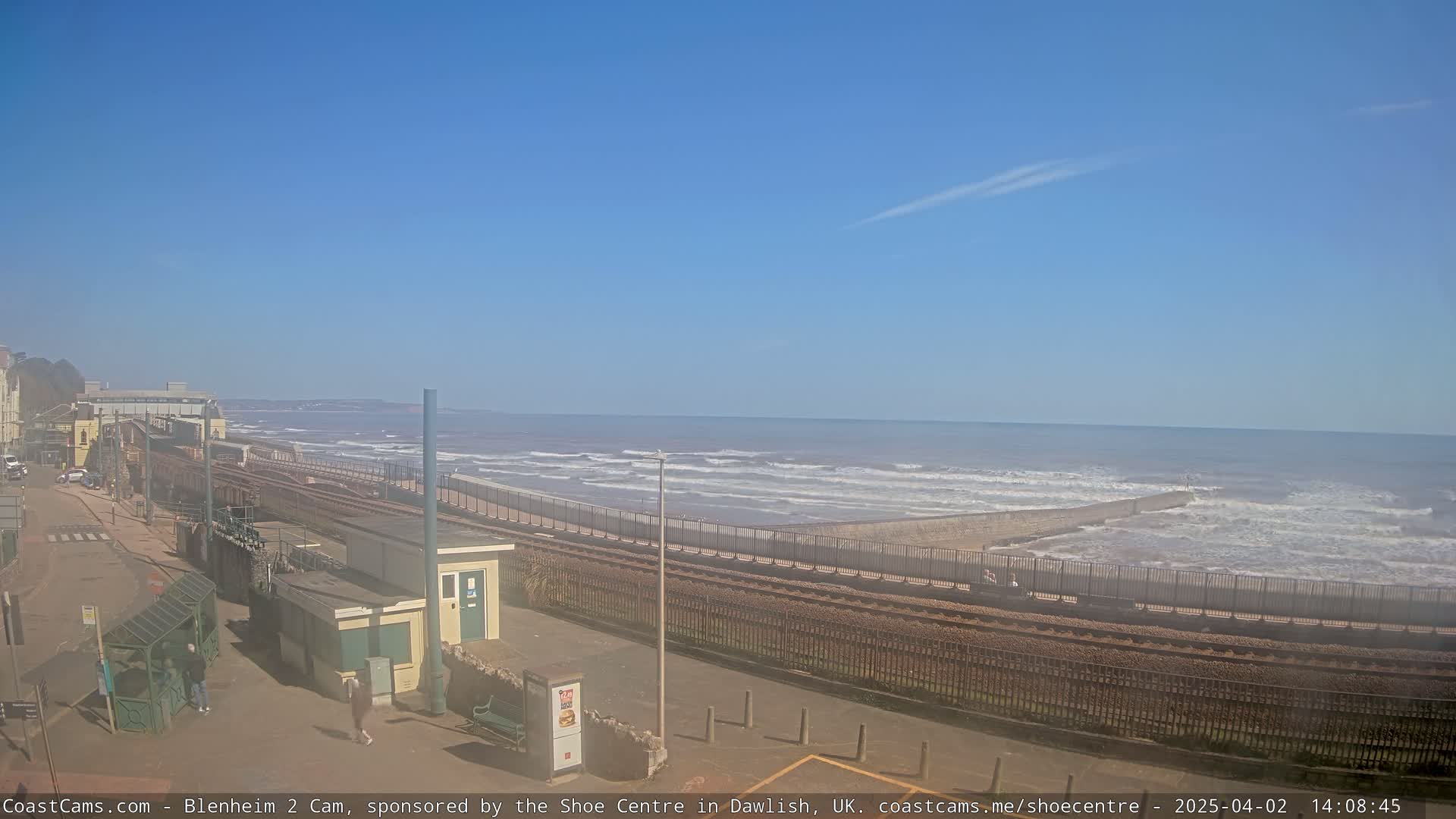 A seaside town scene with a train track running alongside a sea wall, overlooking a wavy ocean under a clear blue sky.