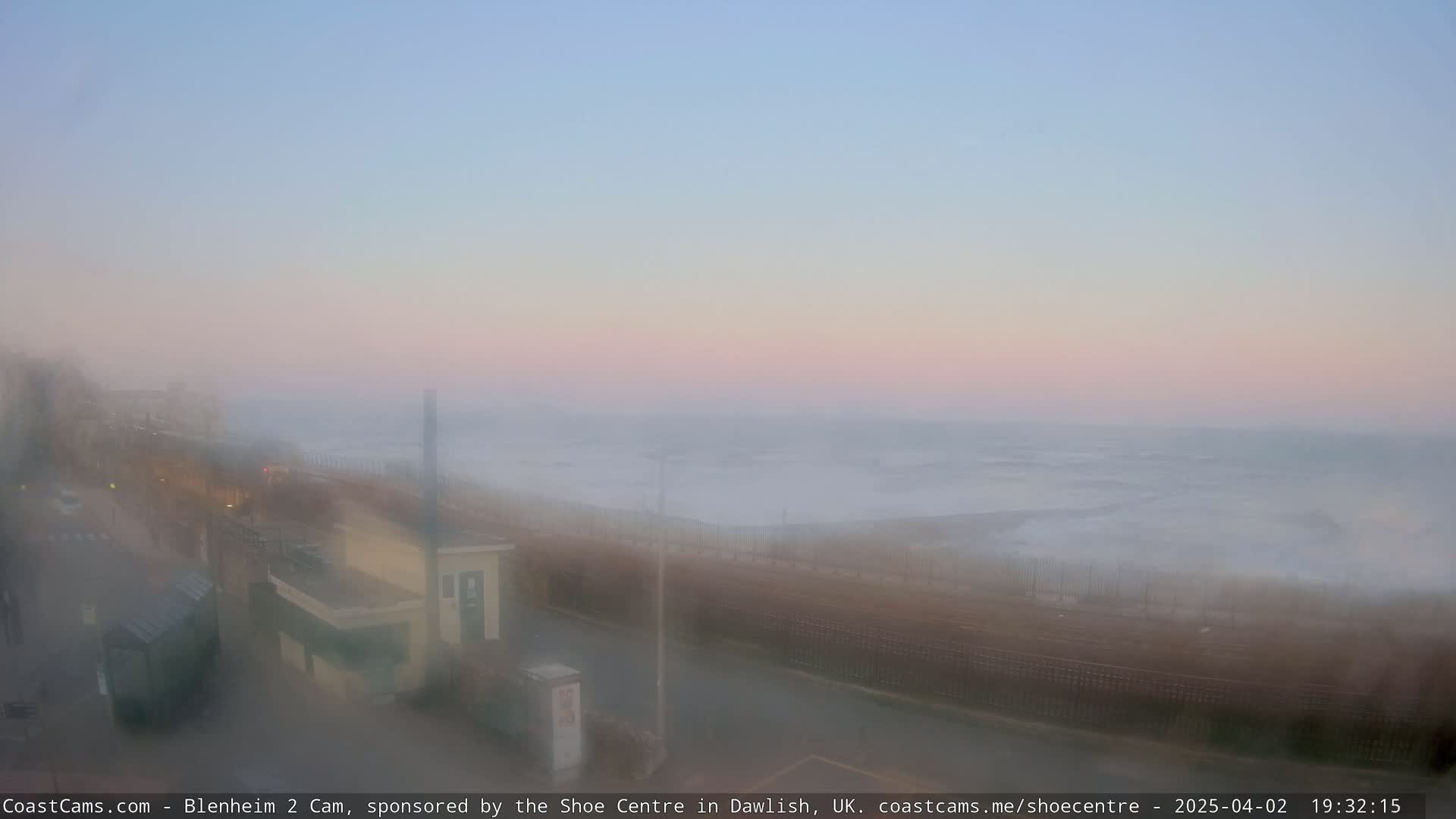 A hazy, pink-tinged sunset over a foggy ocean, viewed from a seaside boardwalk with buildings and a road in the foreground.