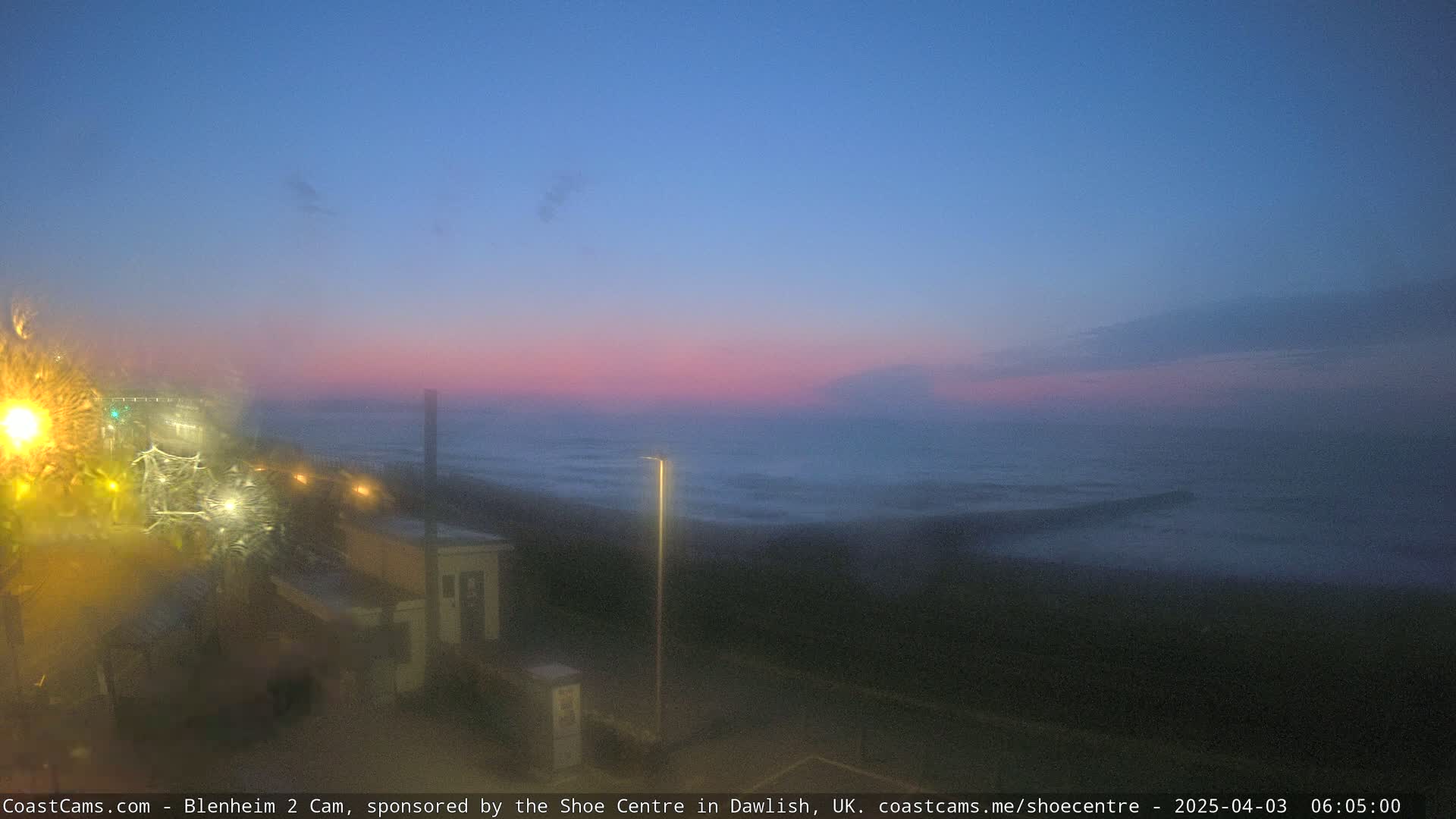 A hazy, pinkish-blue sunrise over a dark, wavy ocean is viewed from a rain-streaked window overlooking a coastal road with lights.