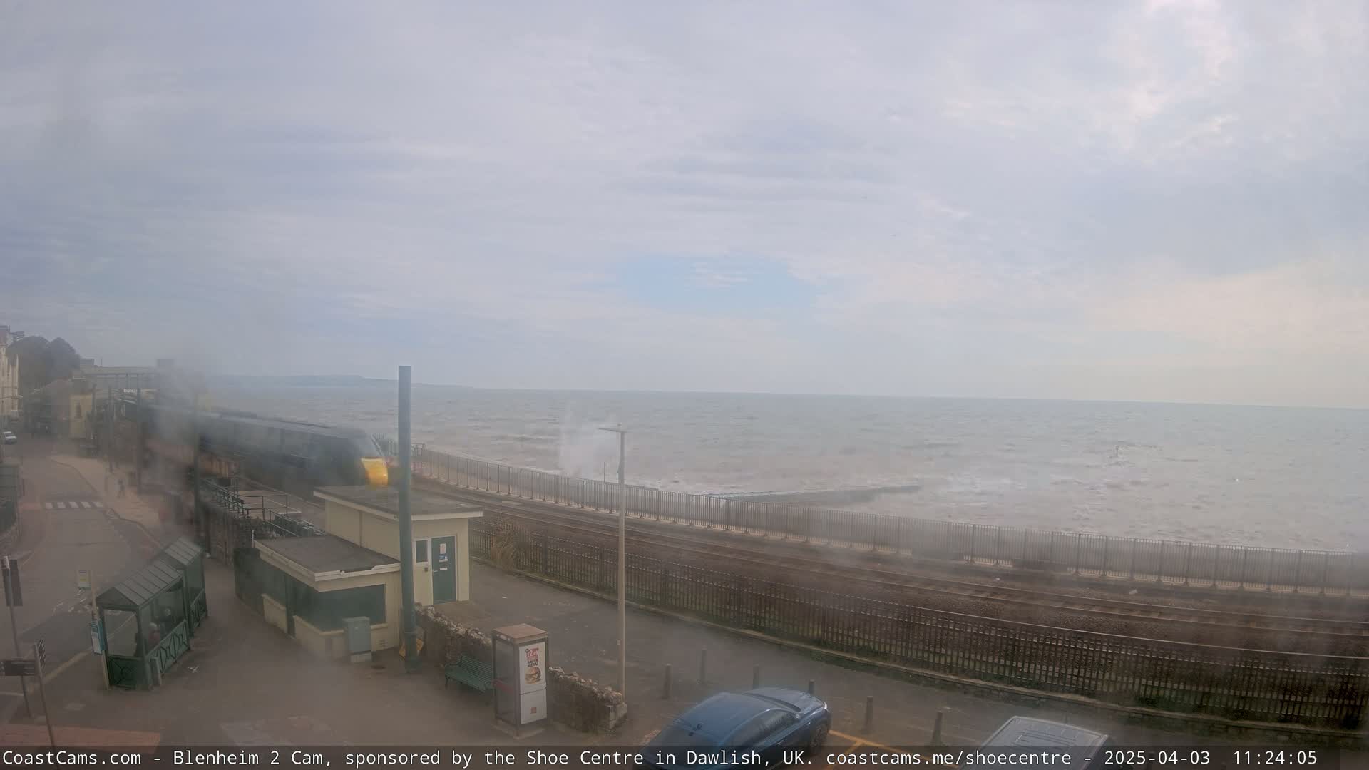 A train passes by a seaside train station on an overcast day with choppy waves crashing against a seawall.