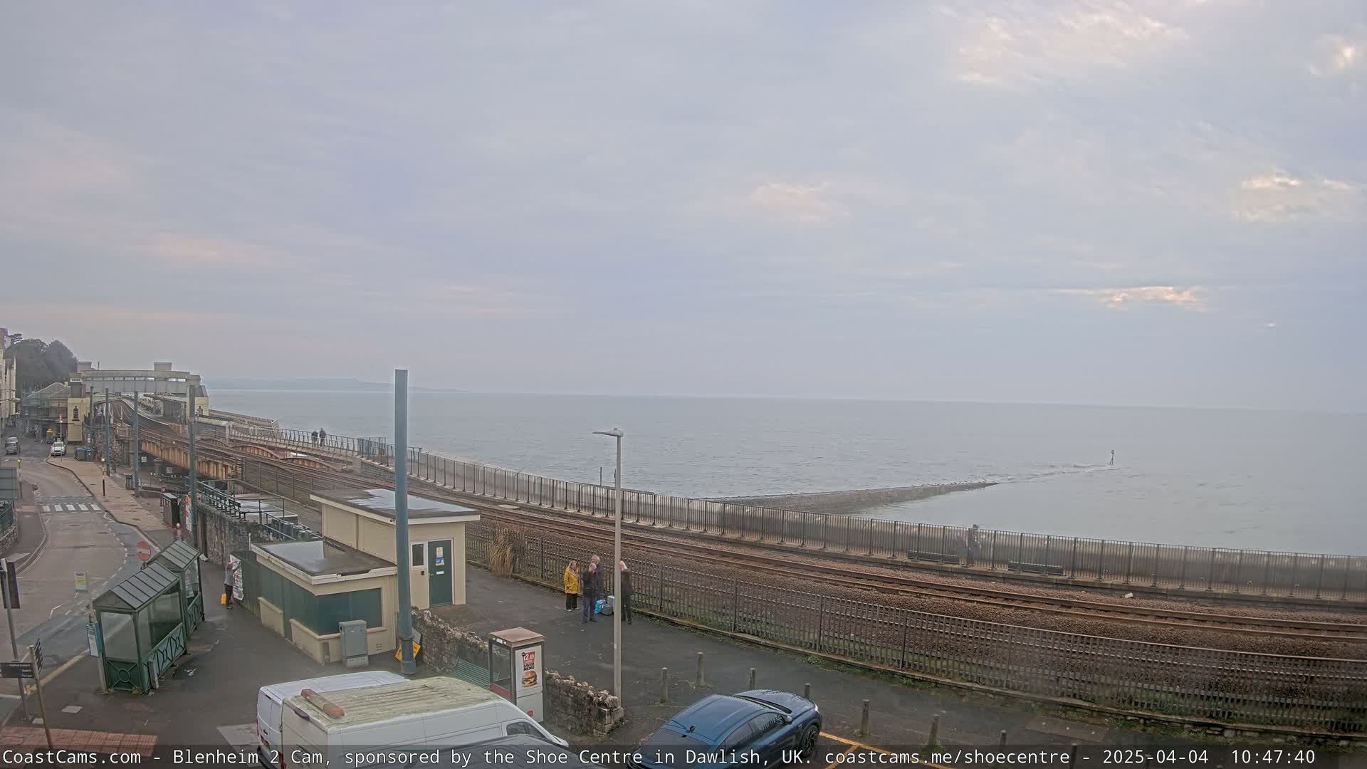 A mostly cloudy day reveals a coastal scene with train tracks running alongside a seawall that extends into a calm sea.
