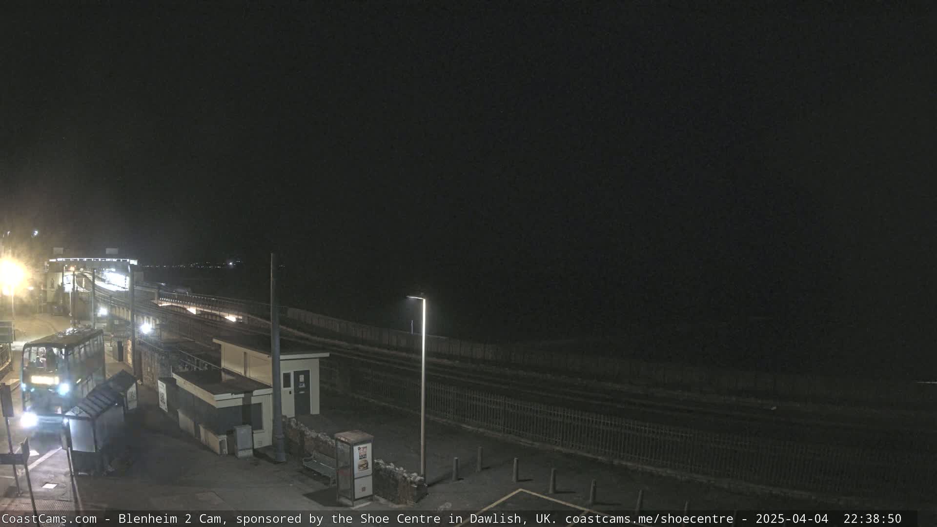 A nighttime view of a train station platform with a bus and a dimly lit area beyond a fence.