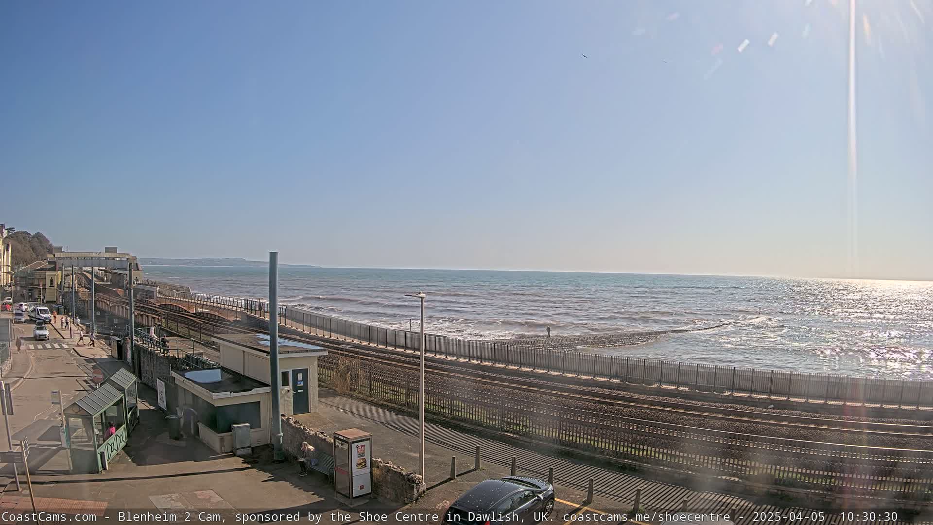 A sunny day shows a coastal train line next to a sea wall with moderate waves crashing against it.