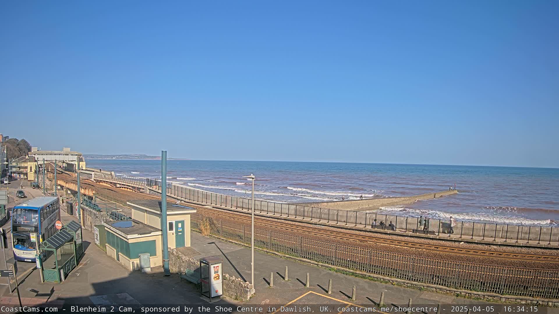 A train track runs alongside a coastline with a calm sea under a clear blue sky, where people are walking along the shore near a low seawall.