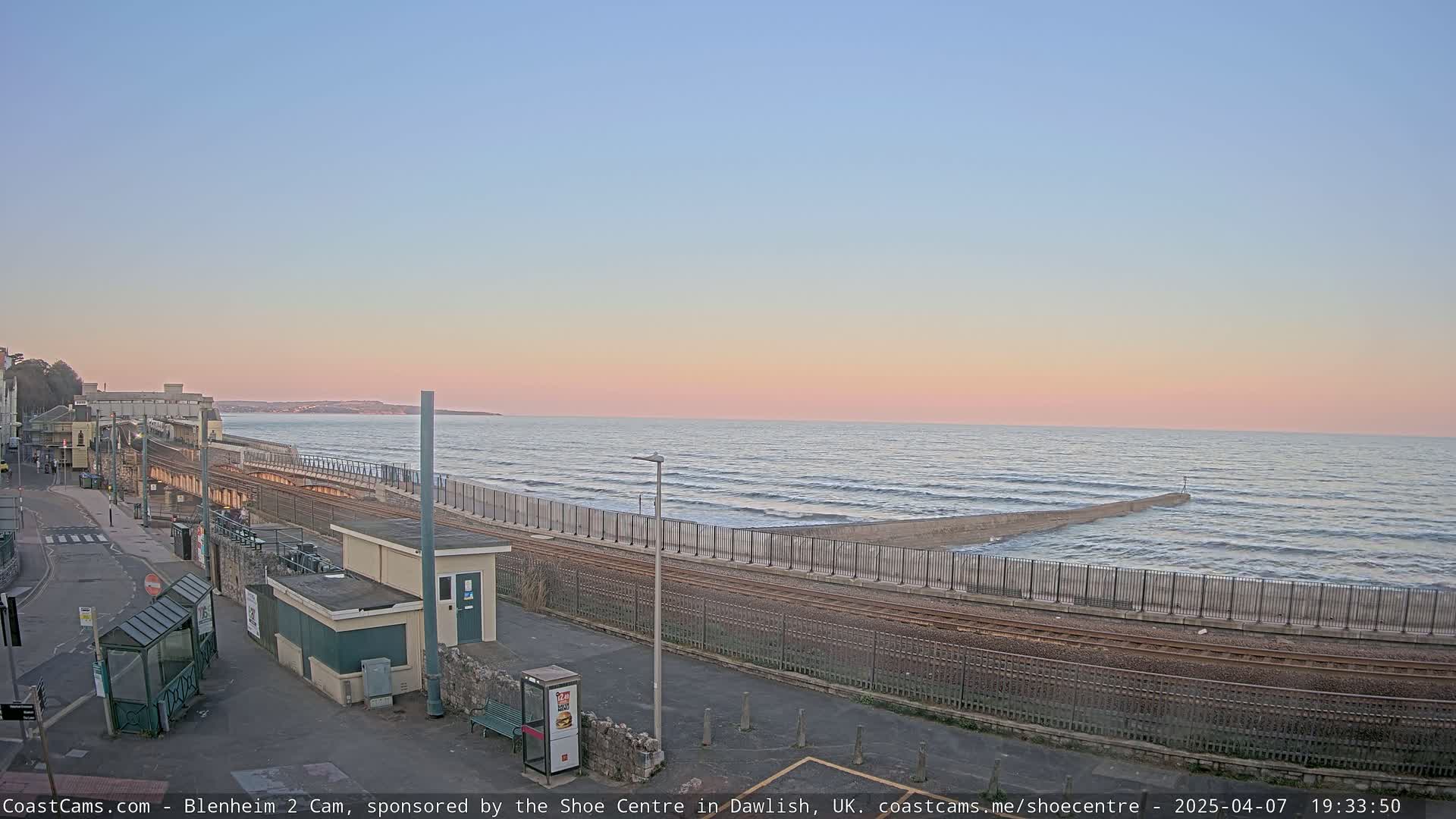 A coastal train track runs alongside a sea wall, with gentle waves lapping the shore under a pastel sunset sky.