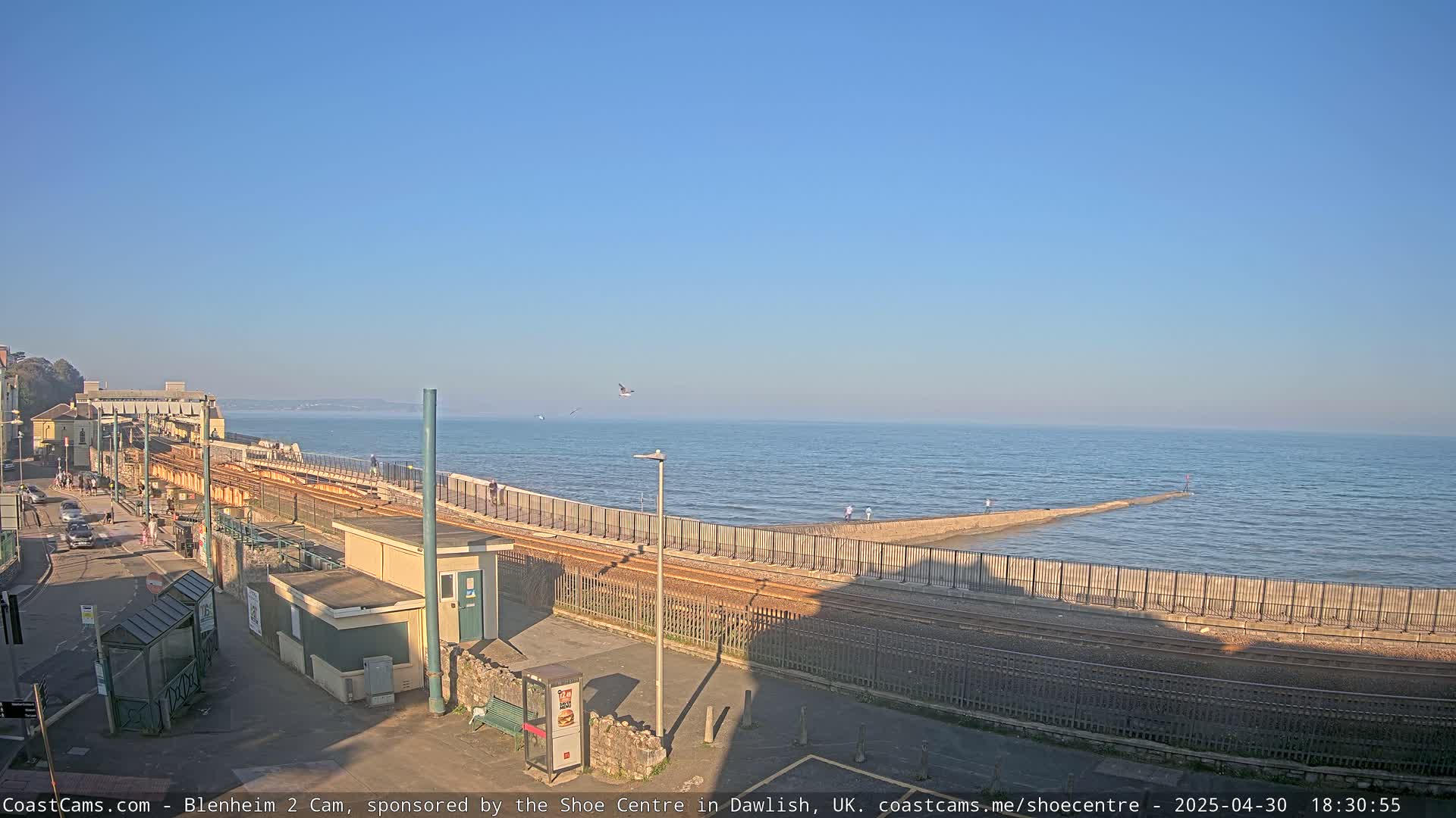 A seaside train track runs alongside a seawall, with calm ocean waters extending to the horizon under a clear blue sky.