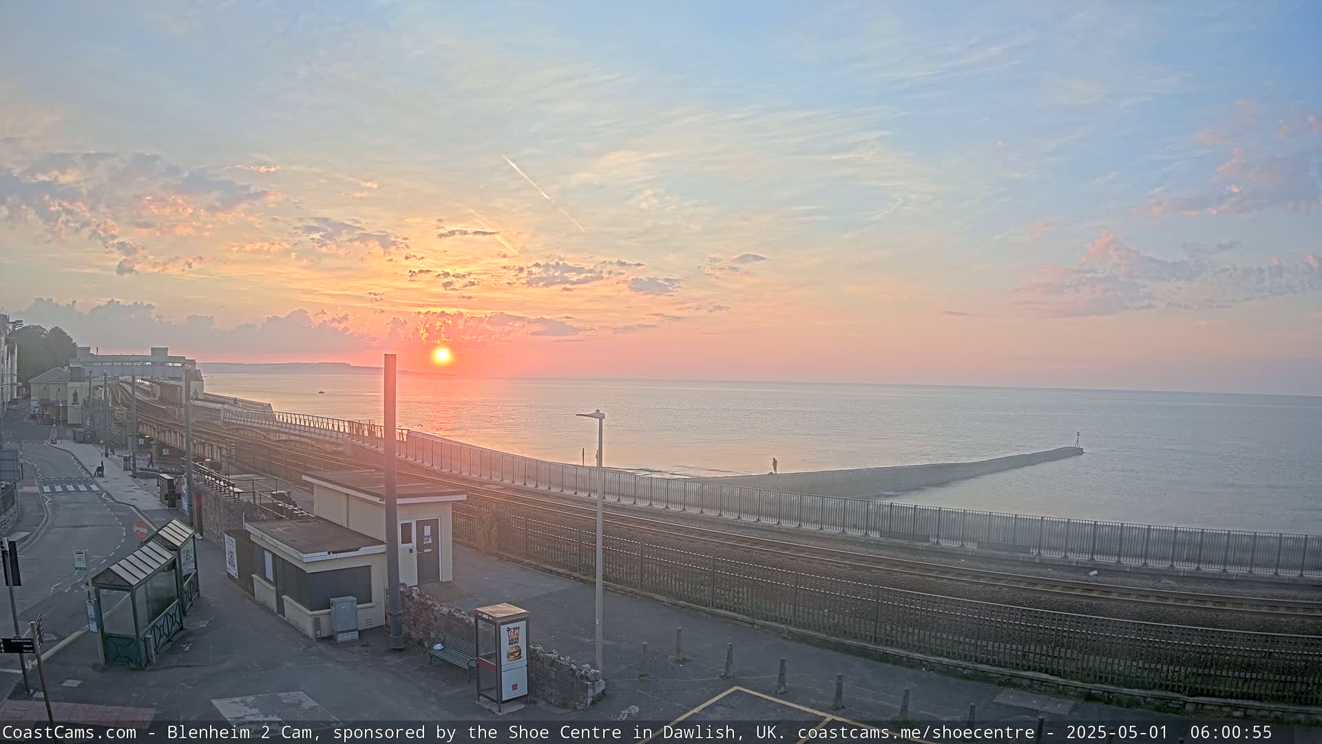 A coastal sunrise scene with calm waters, a railway line bordering the shore, and a small building near the tracks.