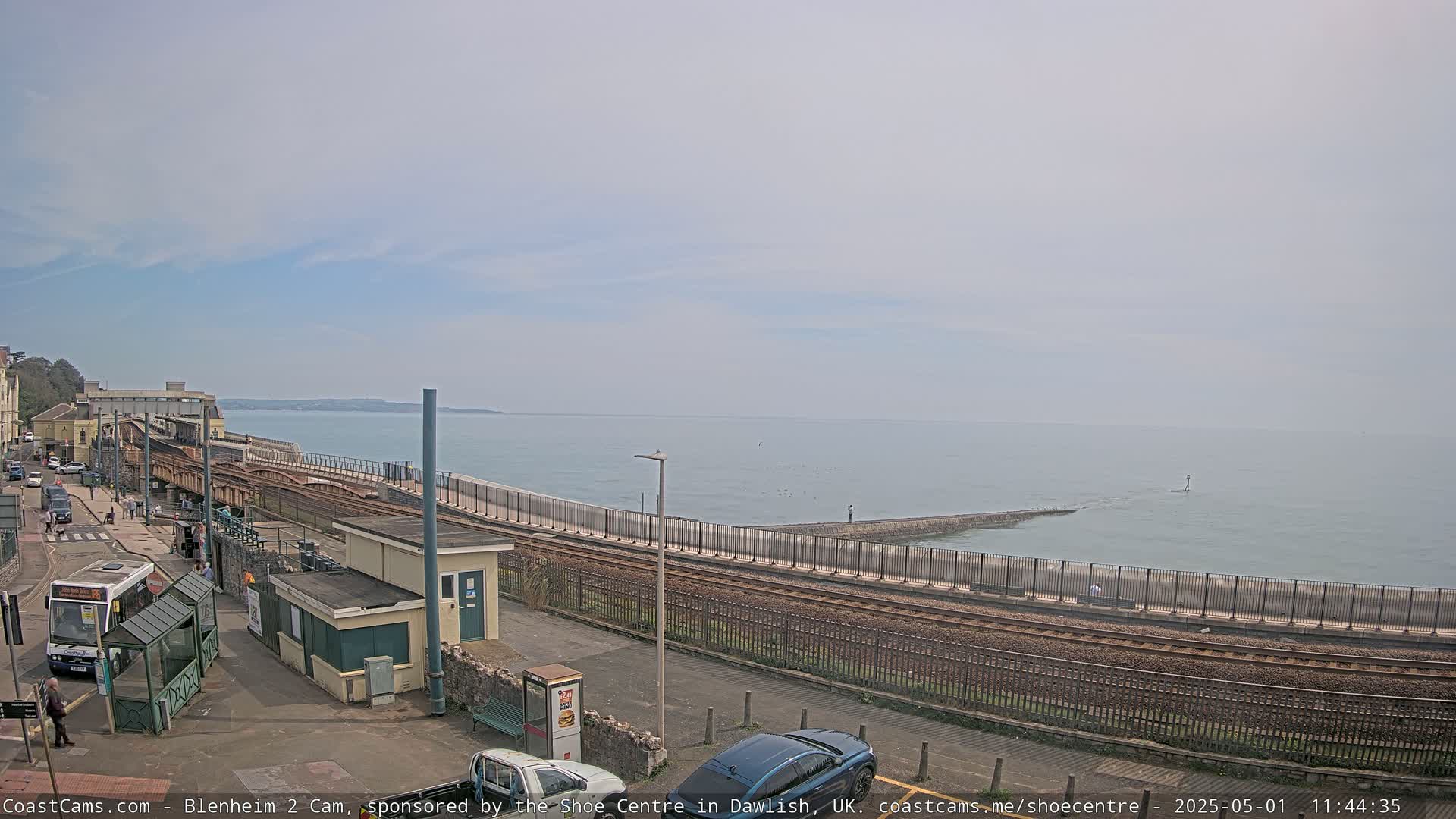 A coastal train track runs alongside a seawall, with a bus stop and cars visible near the tracks under a mostly clear sky.