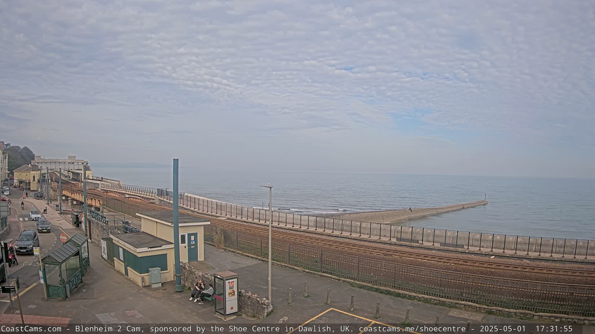 A coastal train track runs alongside a seawall, curving towards a breakwater extending into a calm sea under a mostly cloudy sky.