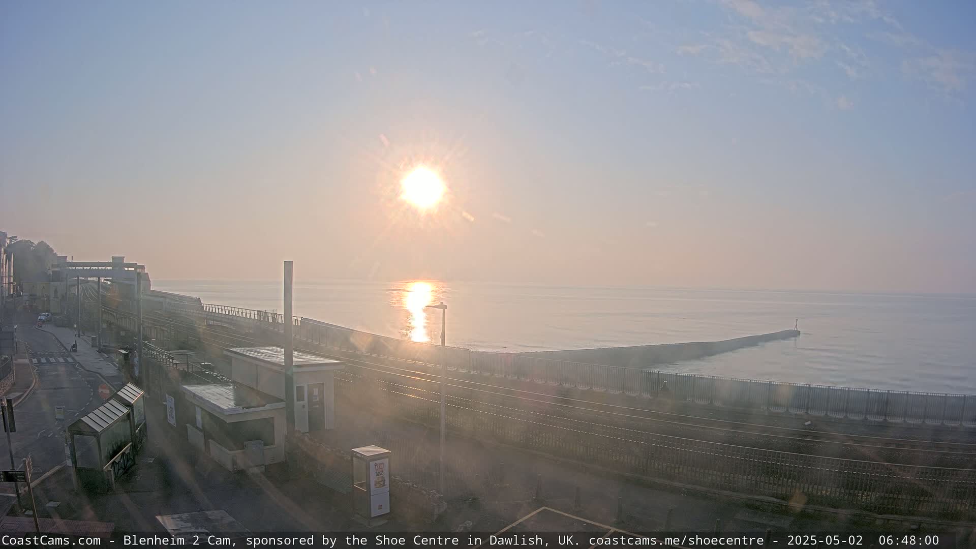 A hazy sunrise illuminates a calm sea, reflecting the sun's light, beside a train track and coastal structures.