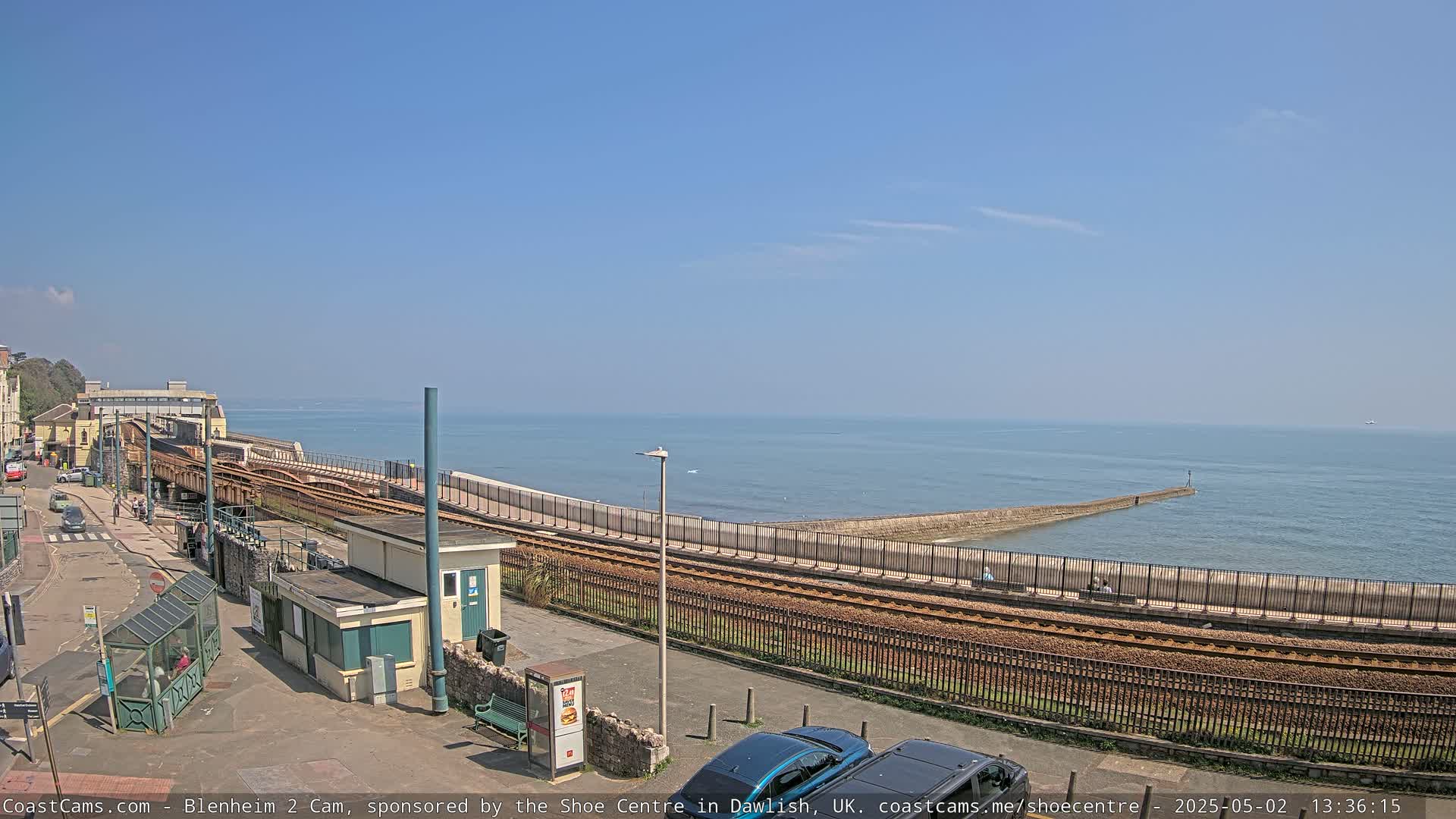 A seaside train track curves along a seawall, extending toward a calm ocean under a clear blue sky.