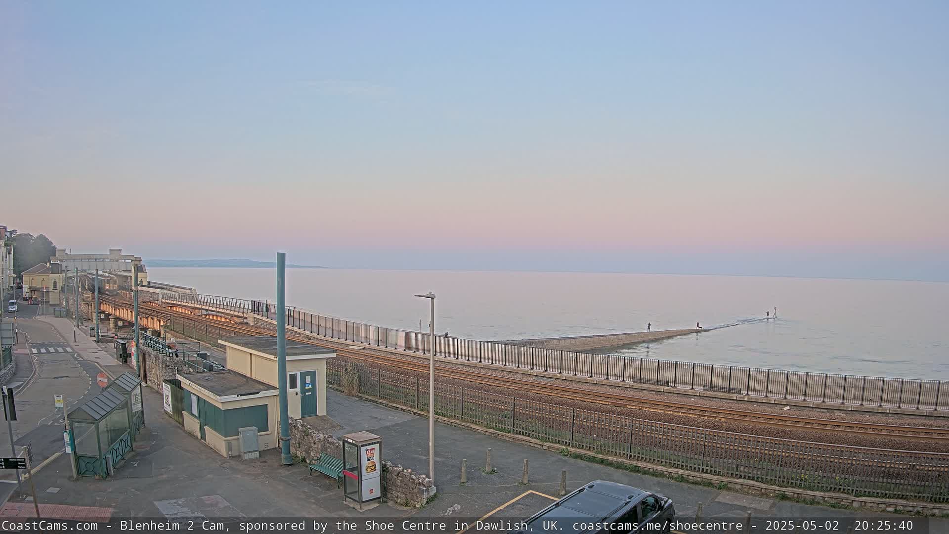 A railway line runs alongside a calm sea under a pale pink and purple sunset.