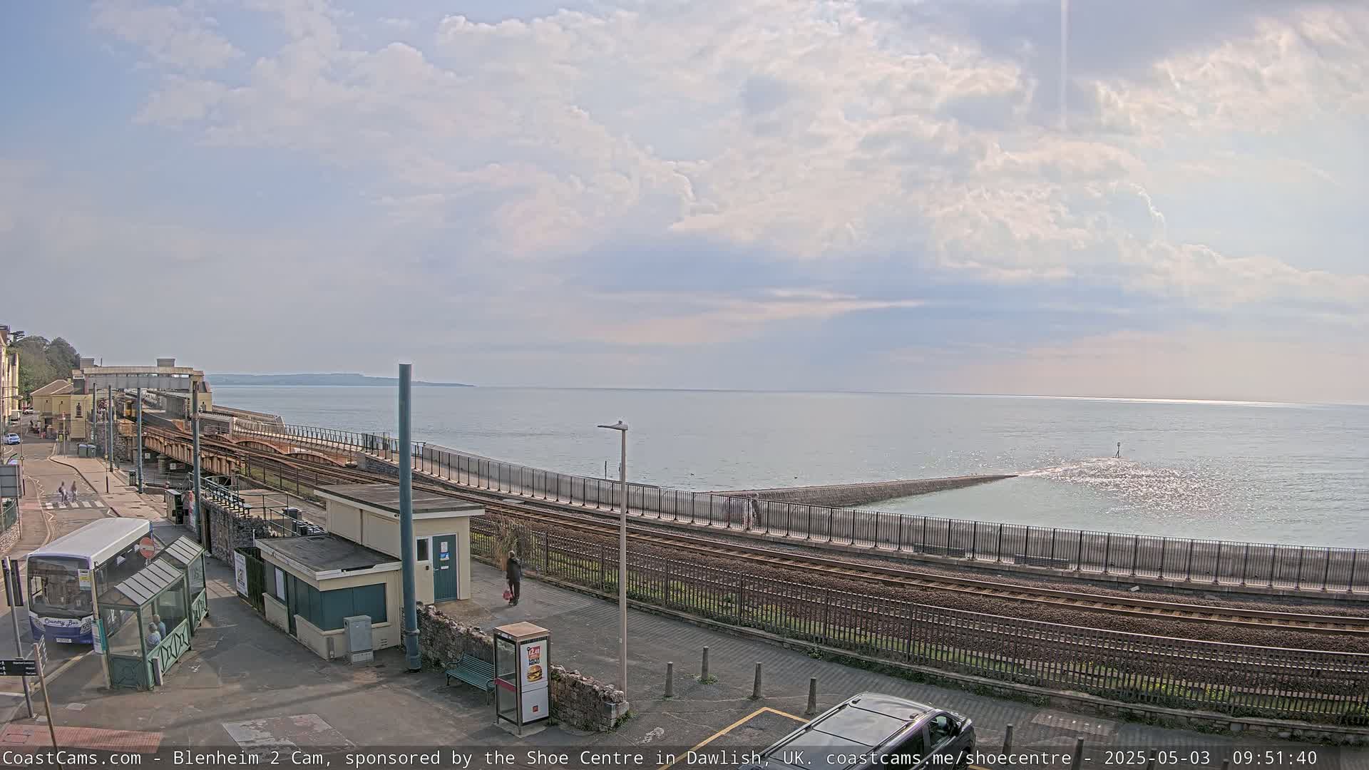 A train track runs alongside a calm ocean under a partly cloudy sky, near a bus stop and small buildings.
