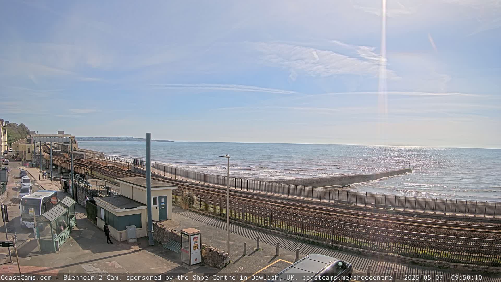 A seaside train track runs alongside a wall separating it from a beach with gently crashing waves under a mostly sunny sky.