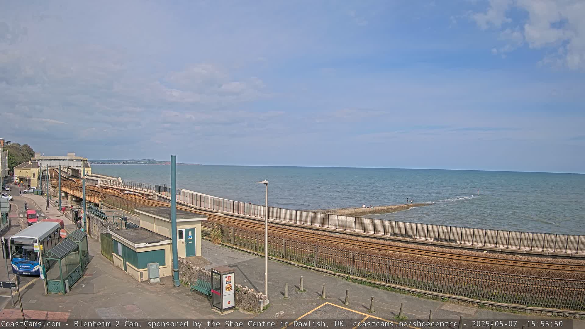A train track runs alongside a sea wall, curving towards a calm ocean under a partly cloudy sky.