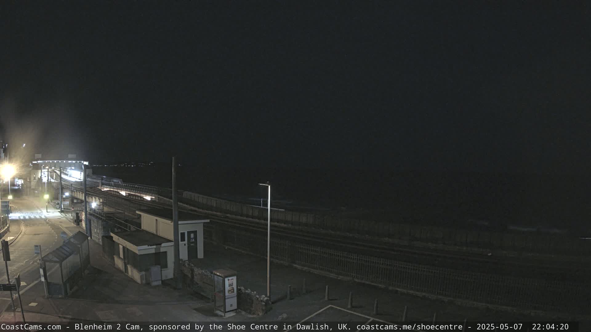 A nighttime view of a coastal train station and tracks, with dark, overcast skies and a calm sea in the distance.