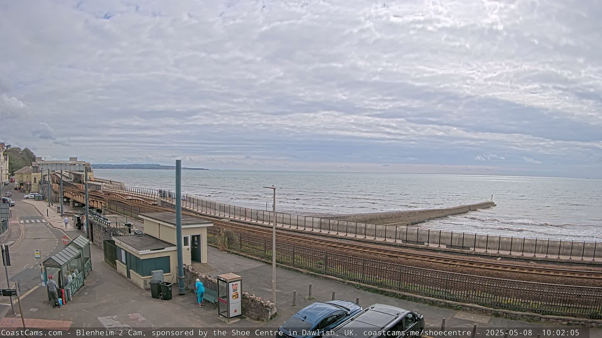 A coastal railway line runs alongside a seawall with moderate waves under a mostly cloudy sky.