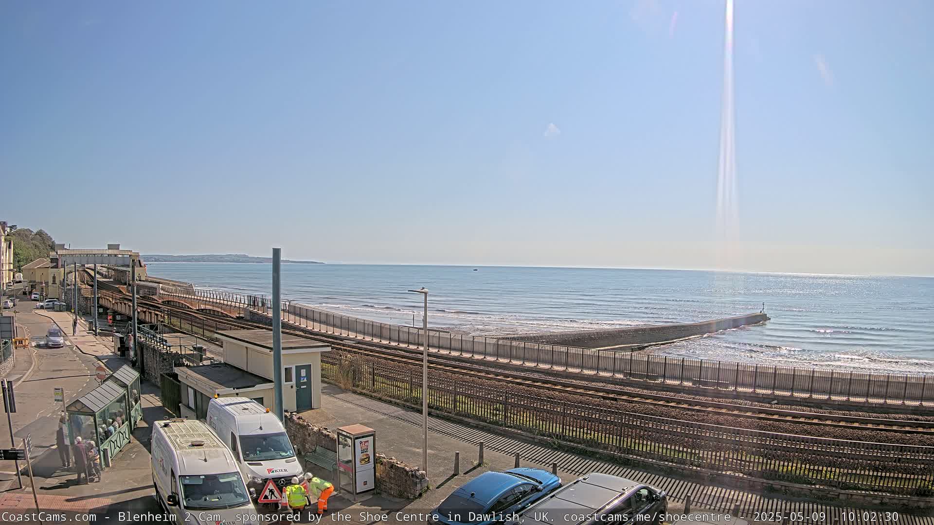 A sunny day at a seaside train station, with calm waves lapping against a seawall and several vehicles parked nearby.