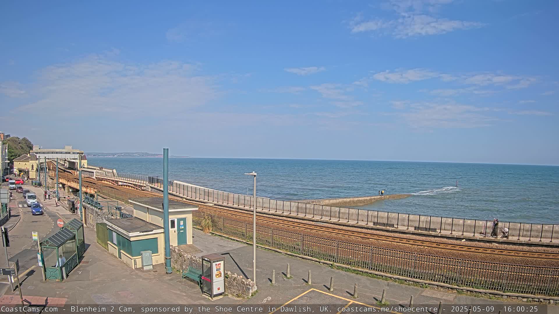 A seaside train track curves alongside a seawall, with a calm ocean and clear blue sky in the background.