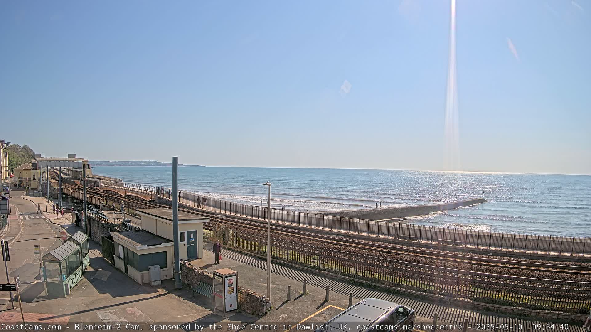A sunny day at a seaside train station shows train tracks running alongside a seawall and a beach with people walking along the shore.