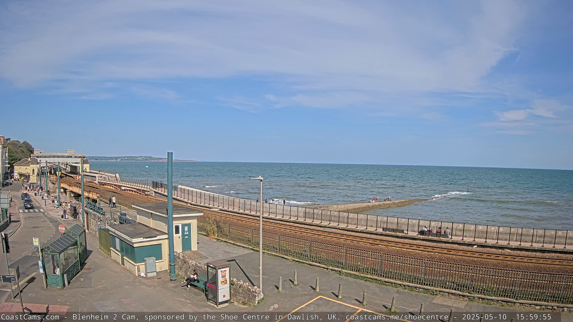A seaside train track runs alongside a calm ocean under a mostly sunny sky, with people walking along the track and shore.
