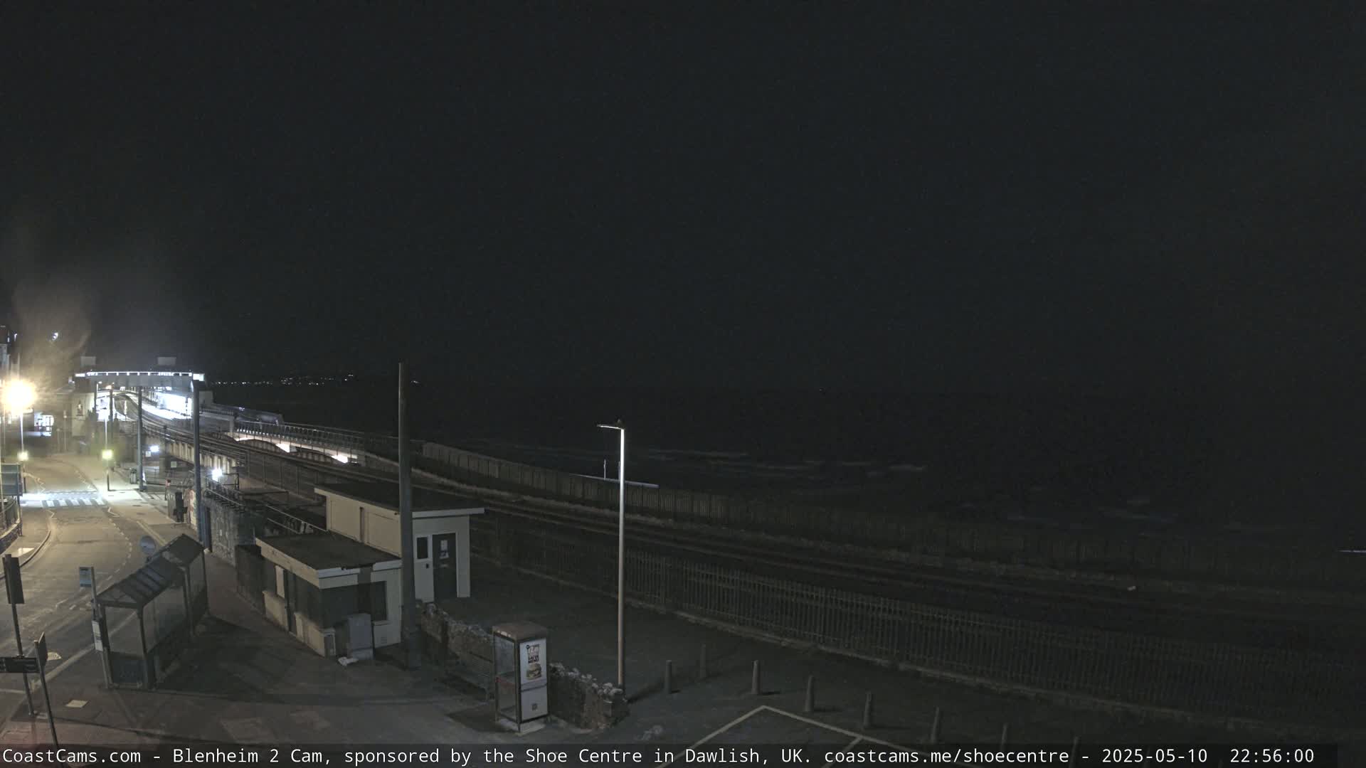 A nighttime coastal scene shows a dark ocean with small waves, a train track running parallel to the shore, and a roadway leading to a building near the water's edge.