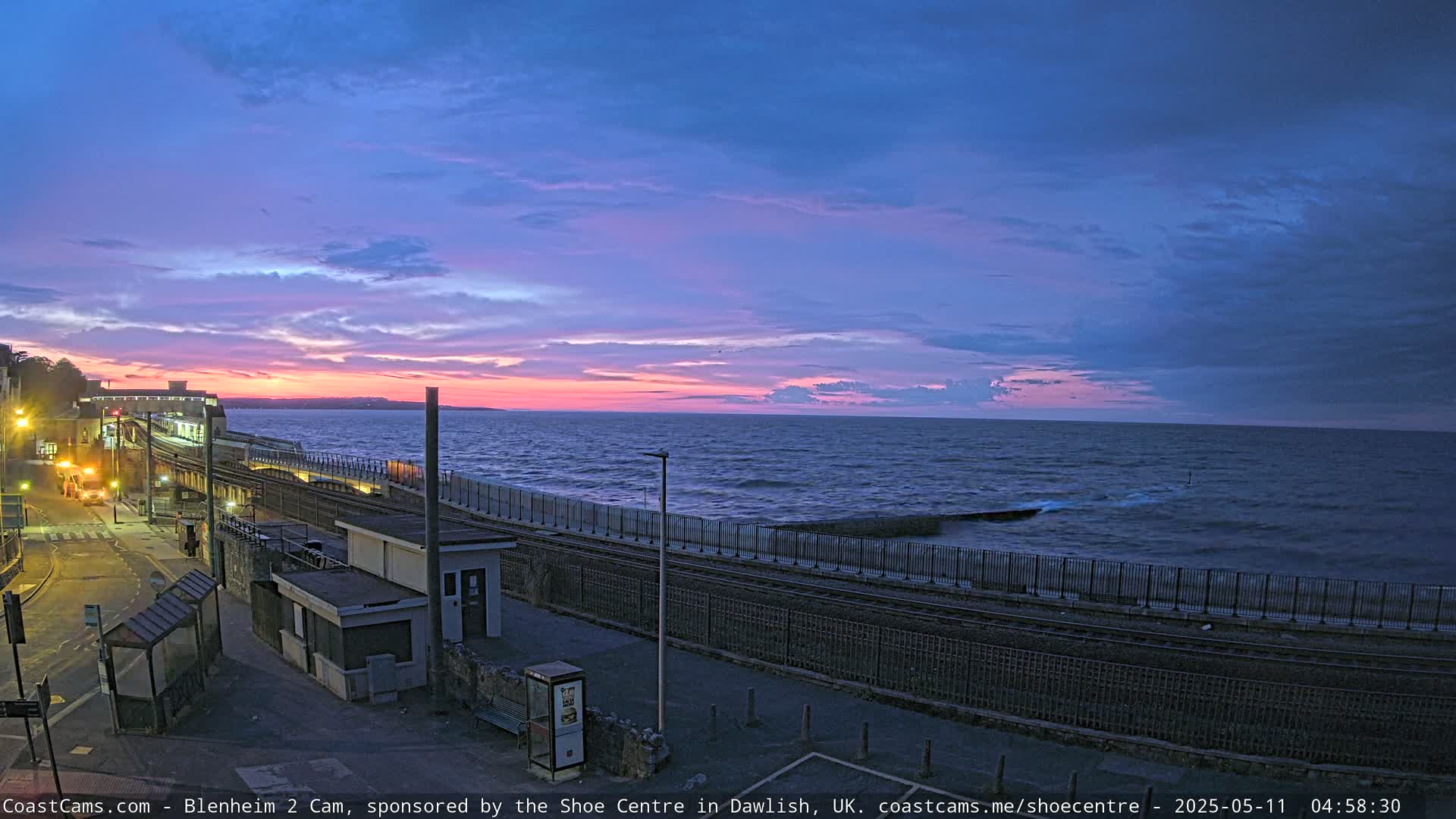 A coastal train track runs alongside a calm sea at sunset under a partly cloudy sky.