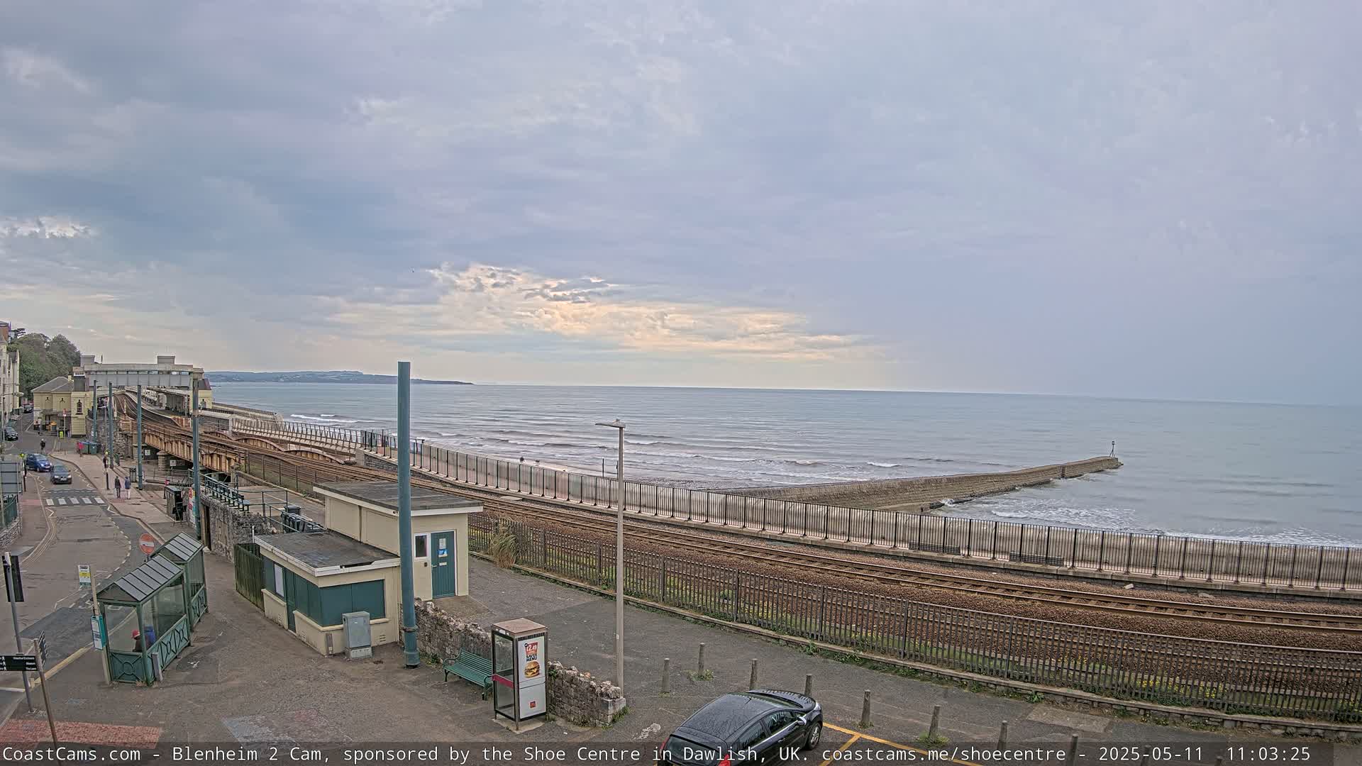 A coastal train station with tracks running alongside a sea wall, overlooking a calm ocean under a cloudy sky.