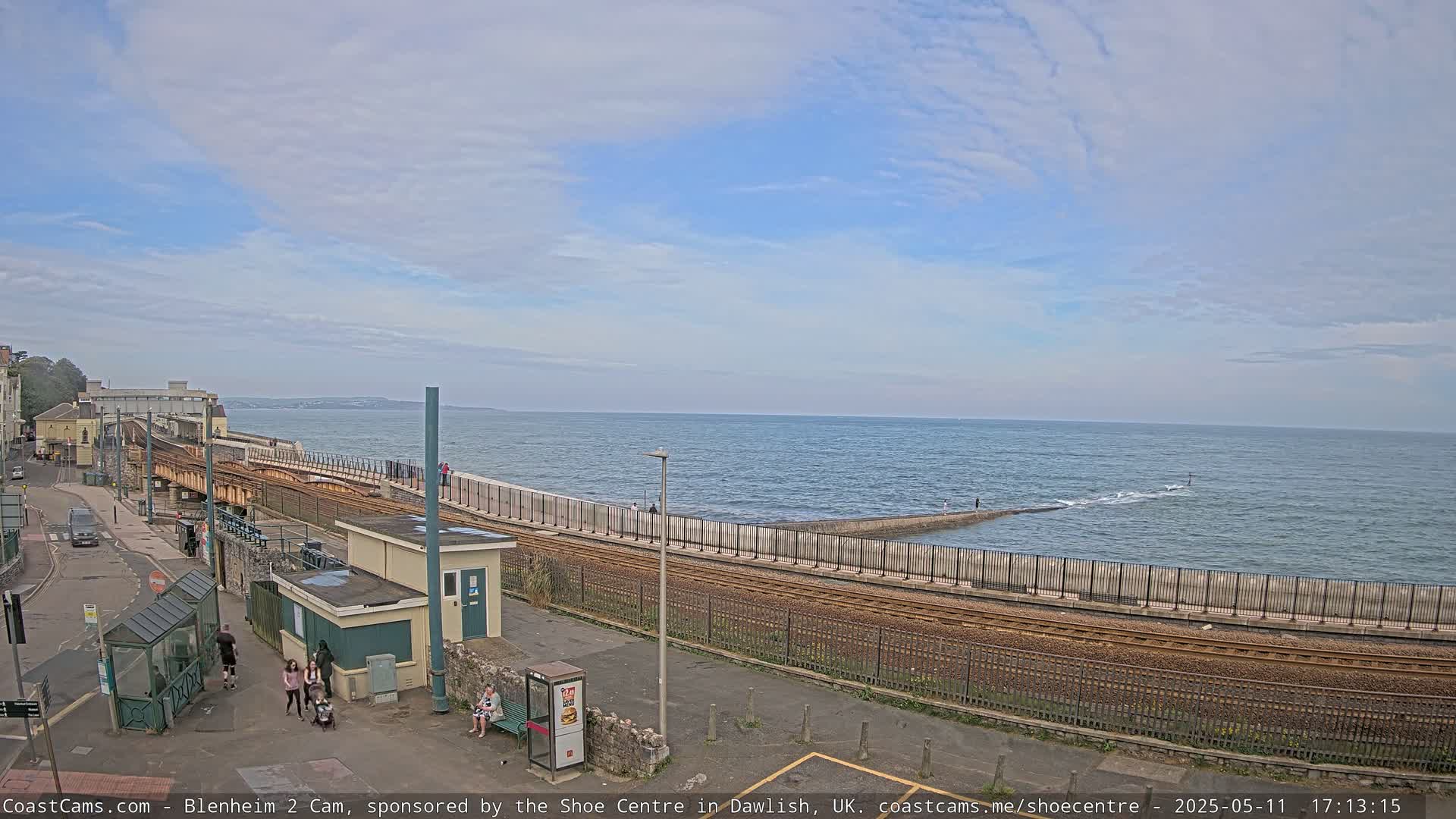 A coastal scene shows a train track curving alongside a seawall, with a calm sea and partly cloudy sky.