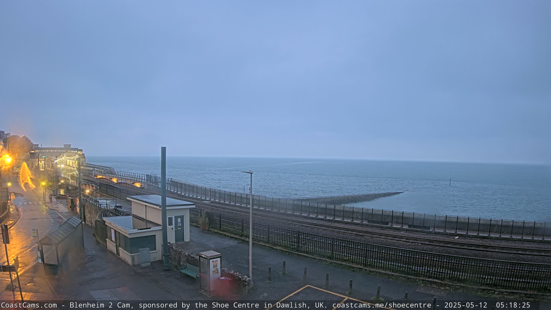 A train station sits beside a calm, gray sea under an overcast sky.