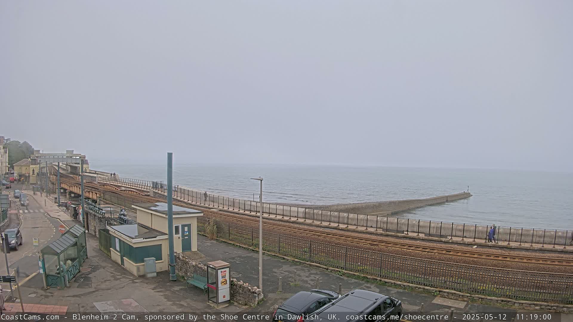 A foggy coastal scene shows a railway line running alongside a seawall next to a calm ocean.