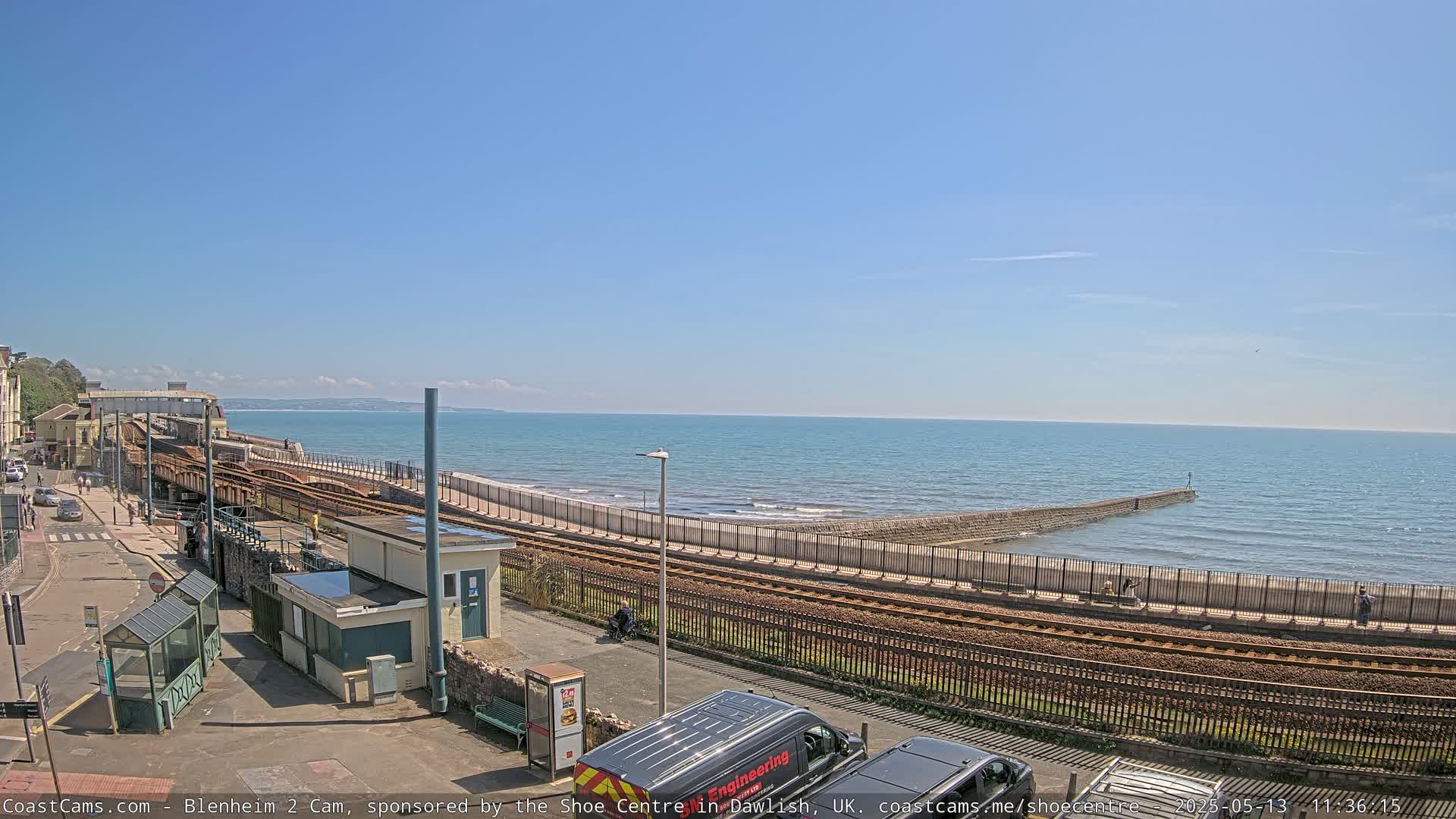 A sunny day at a seaside train station, featuring a calm ocean, a seawall, and railway tracks alongside a paved road.
