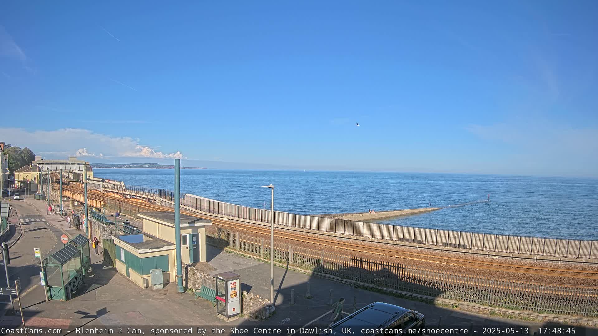 A coastal railway track runs alongside a sea wall, with a calm ocean extending to a distant horizon under a clear blue sky.