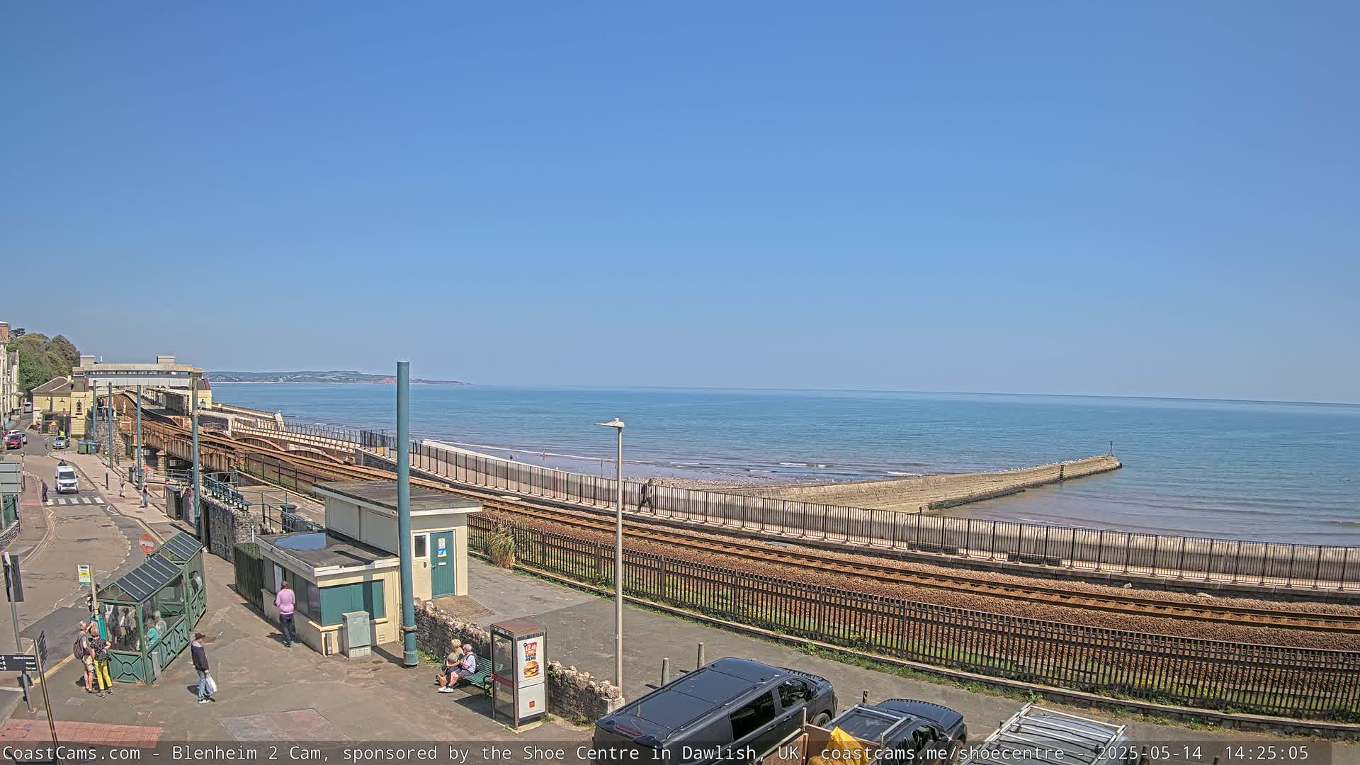 A sunny day at a seaside train station shows train tracks curving alongside a beach with a calm ocean and several people walking nearby.