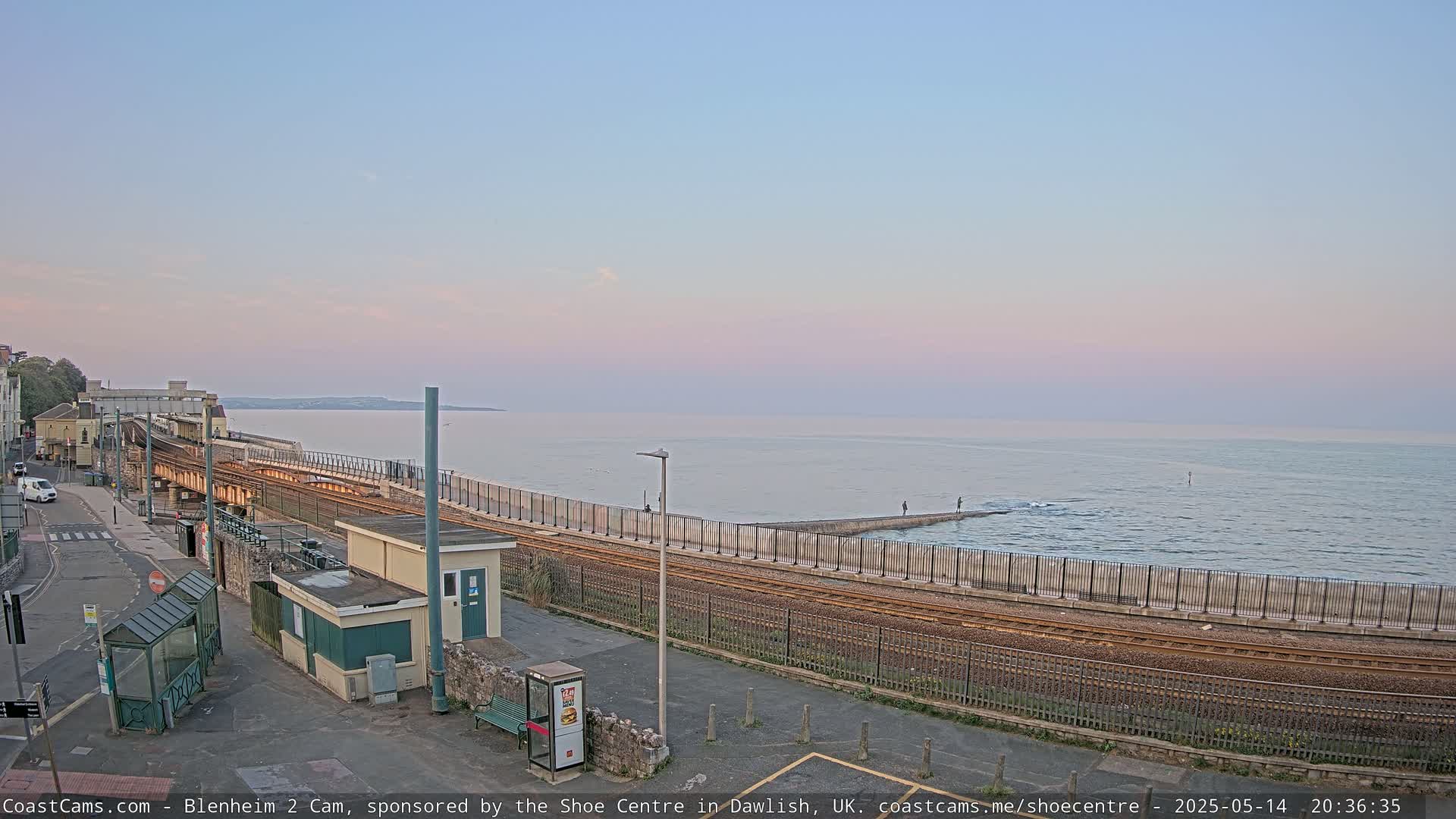 A train track runs alongside a seawall separating a calm ocean under a pale pink and lavender sky at dusk.
