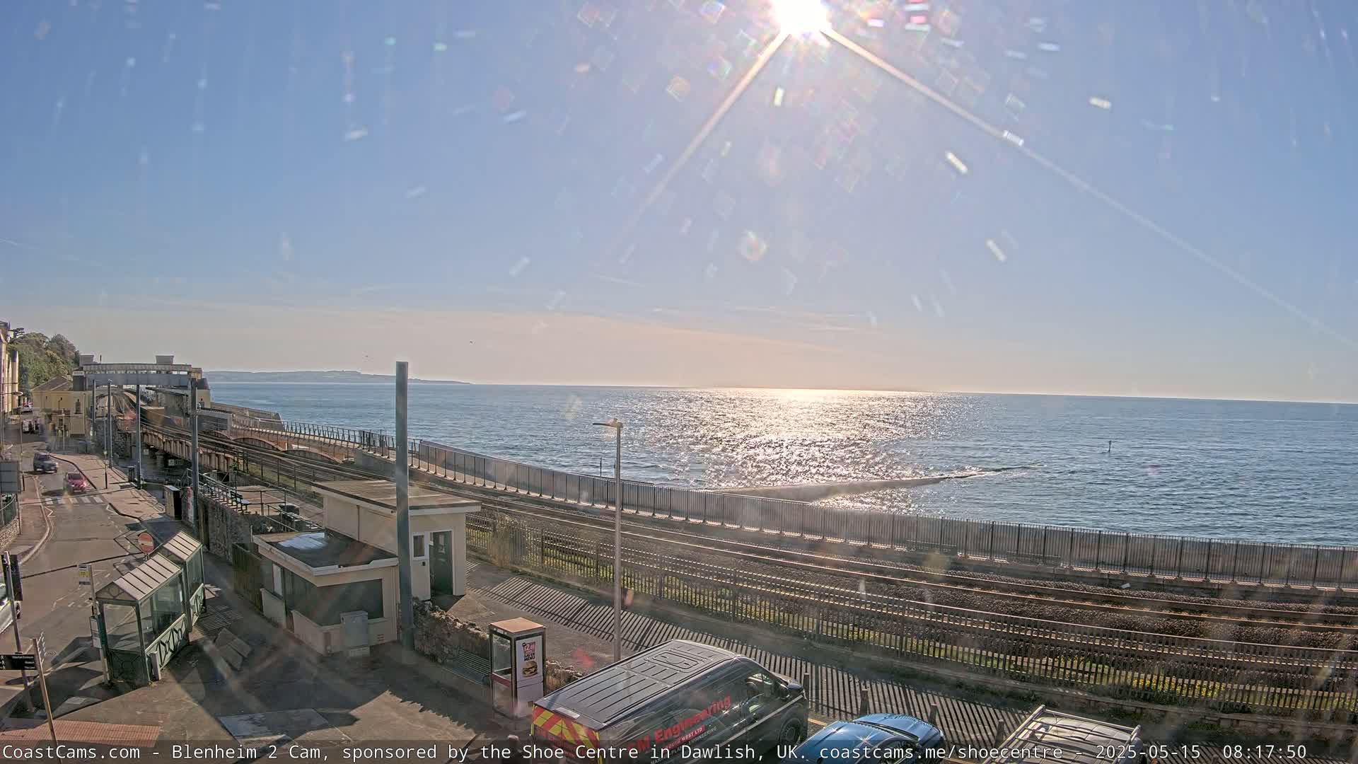 A sunny day shows a coastal railway line next to a calm sea, with a road and buildings nearby.
