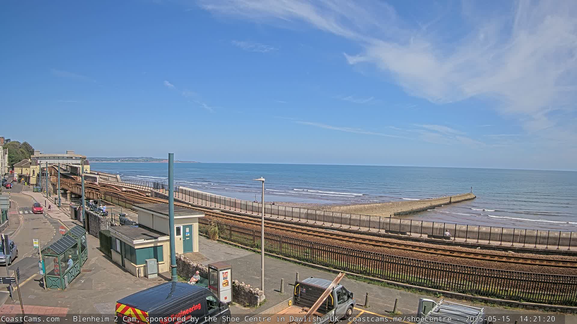 A coastal scene on a partly sunny day shows train tracks running alongside a beach with a calm sea and a low seawall.