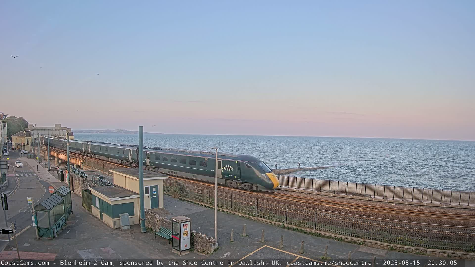 A long, green train travels along a coastal railway line at sunset, with a calm sea visible beyond a low wall.