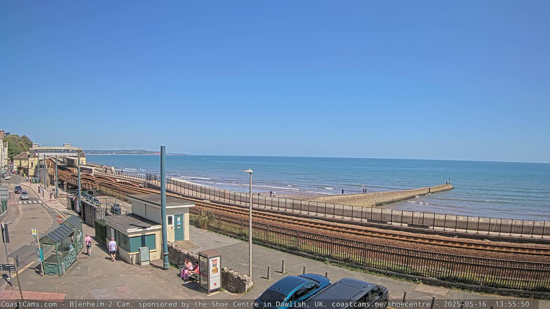 A sunny day shows a coastal town with train tracks running alongside a beach and a calm ocean.