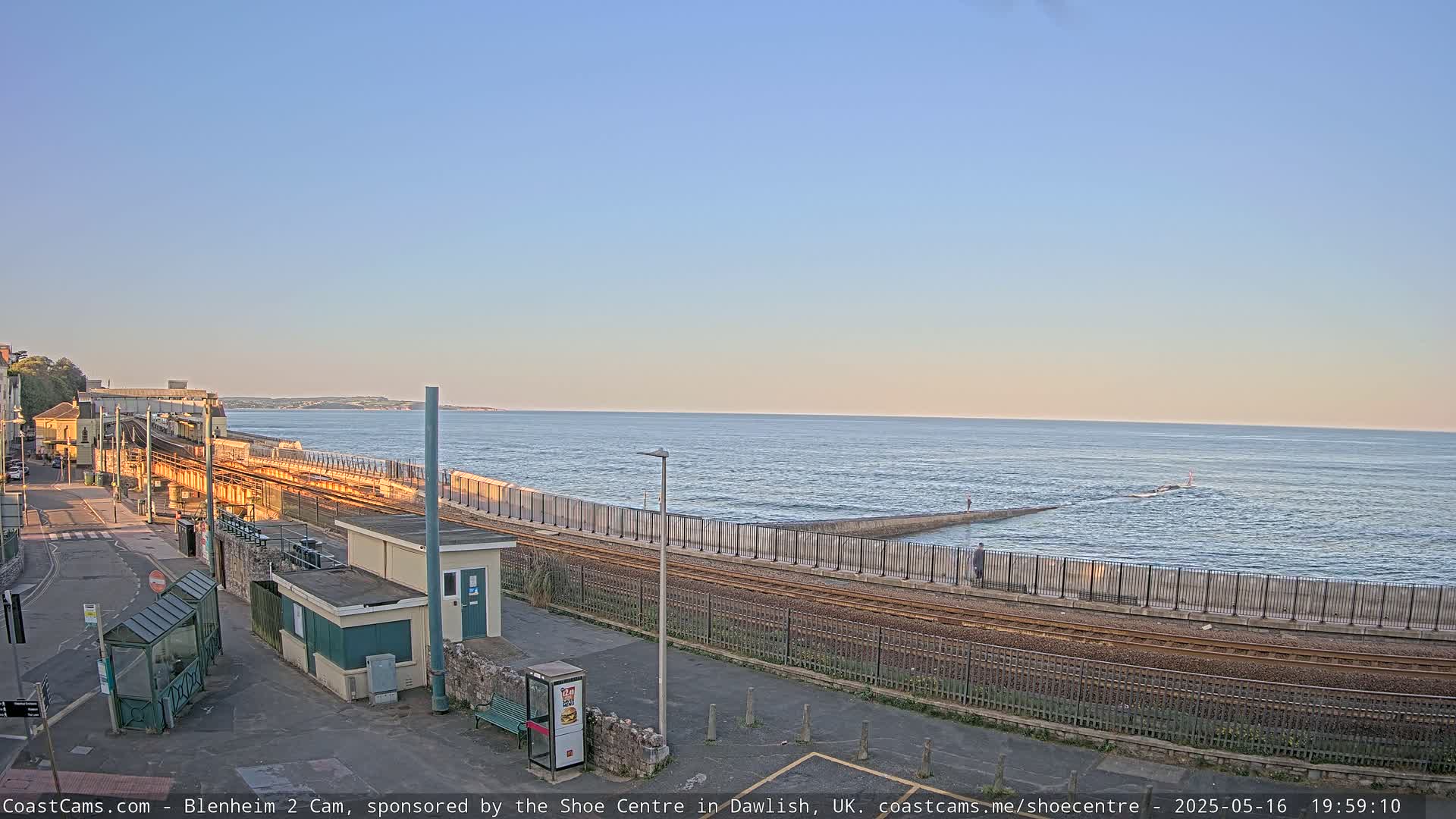 A train track runs alongside a sea wall next to a calm ocean under a clear, pale-orange sunset sky.