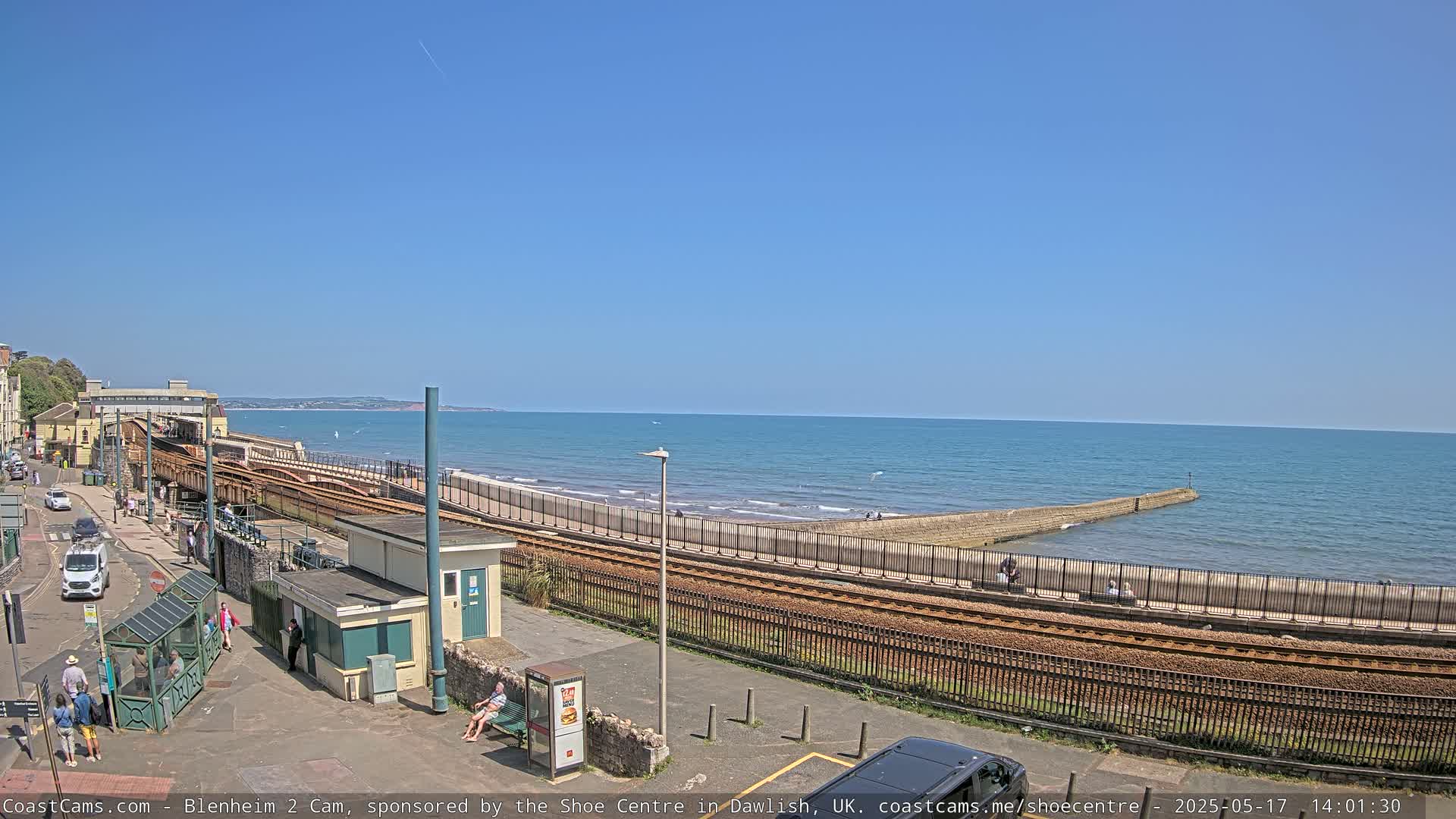 A seaside train track runs parallel to a calm, blue ocean under a clear, sunny sky.