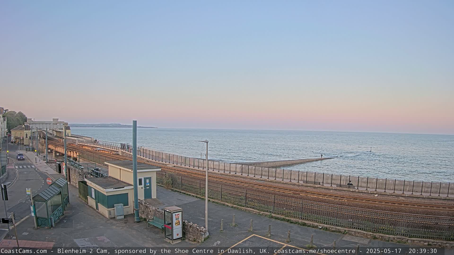 A coastal train track runs alongside a seawall, with a calm sea under a pastel sunset sky.