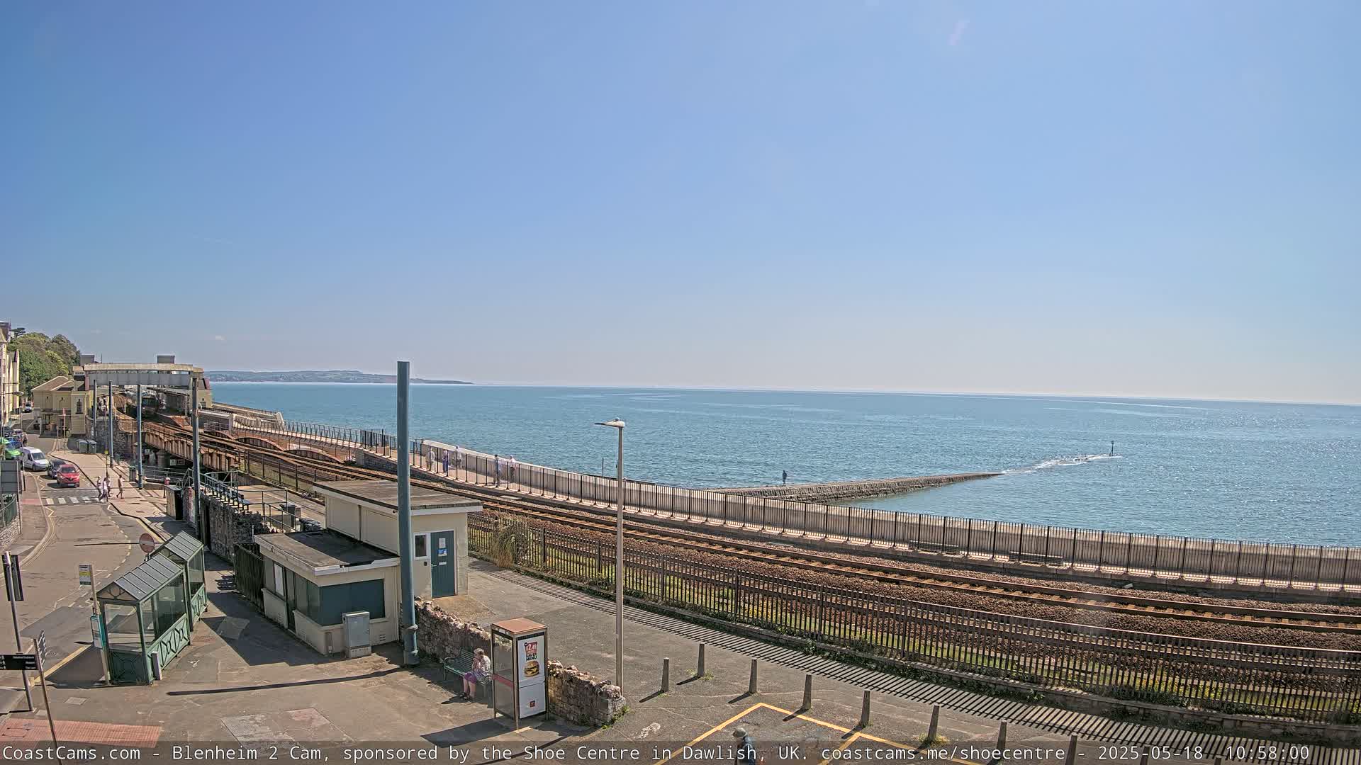 A coastal railway line runs alongside a sea wall, overlooking a calm ocean under a clear blue sky.