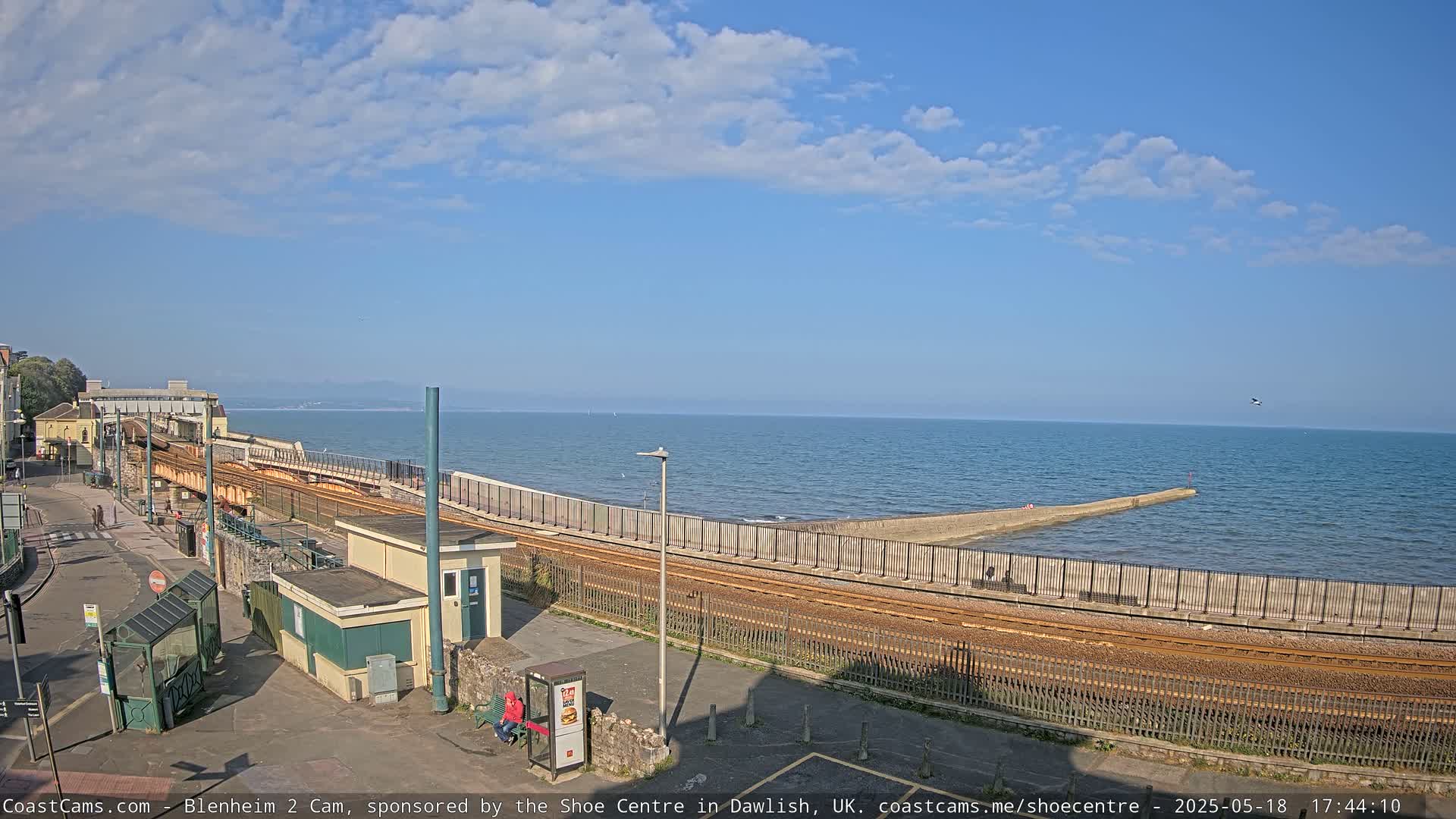 A train track runs alongside a seawall separating a coastal town from a calm ocean under a partly cloudy sky.