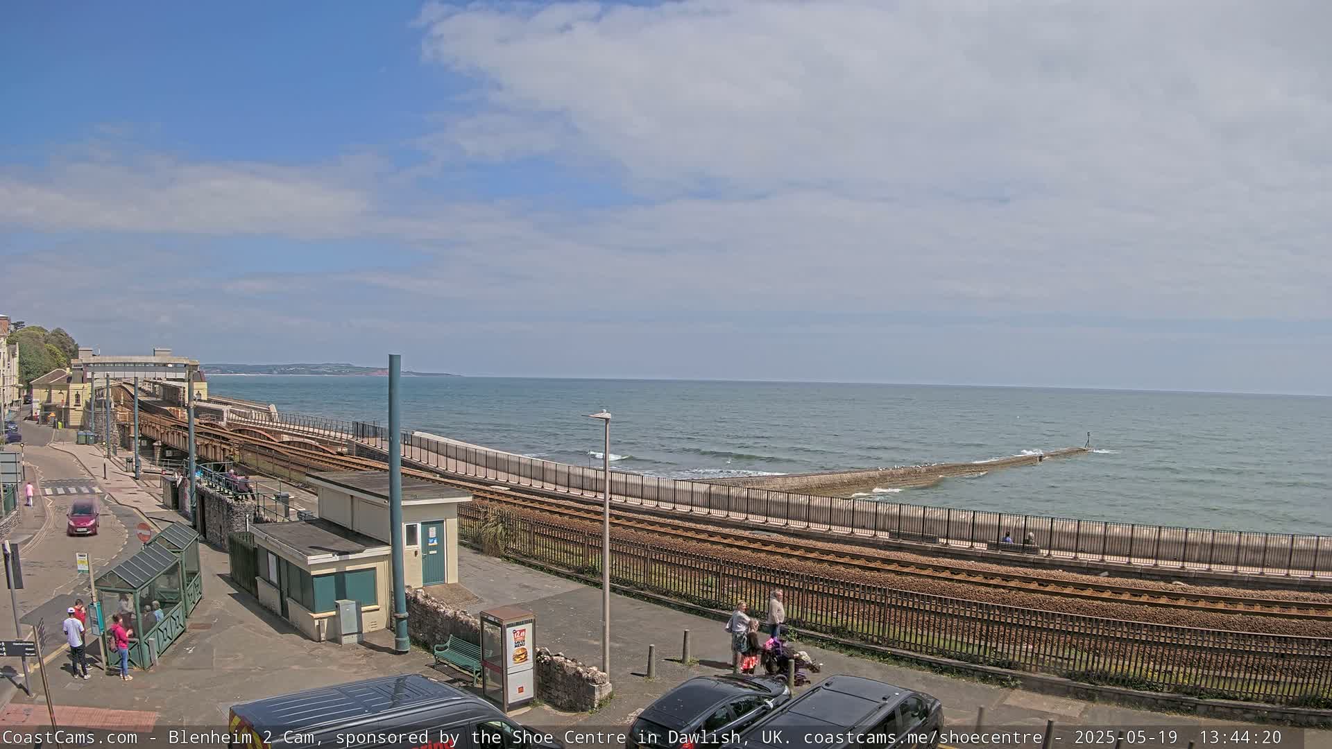 A seaside train track runs alongside a wall bordering a calm sea with small waves under a mostly clear sky.