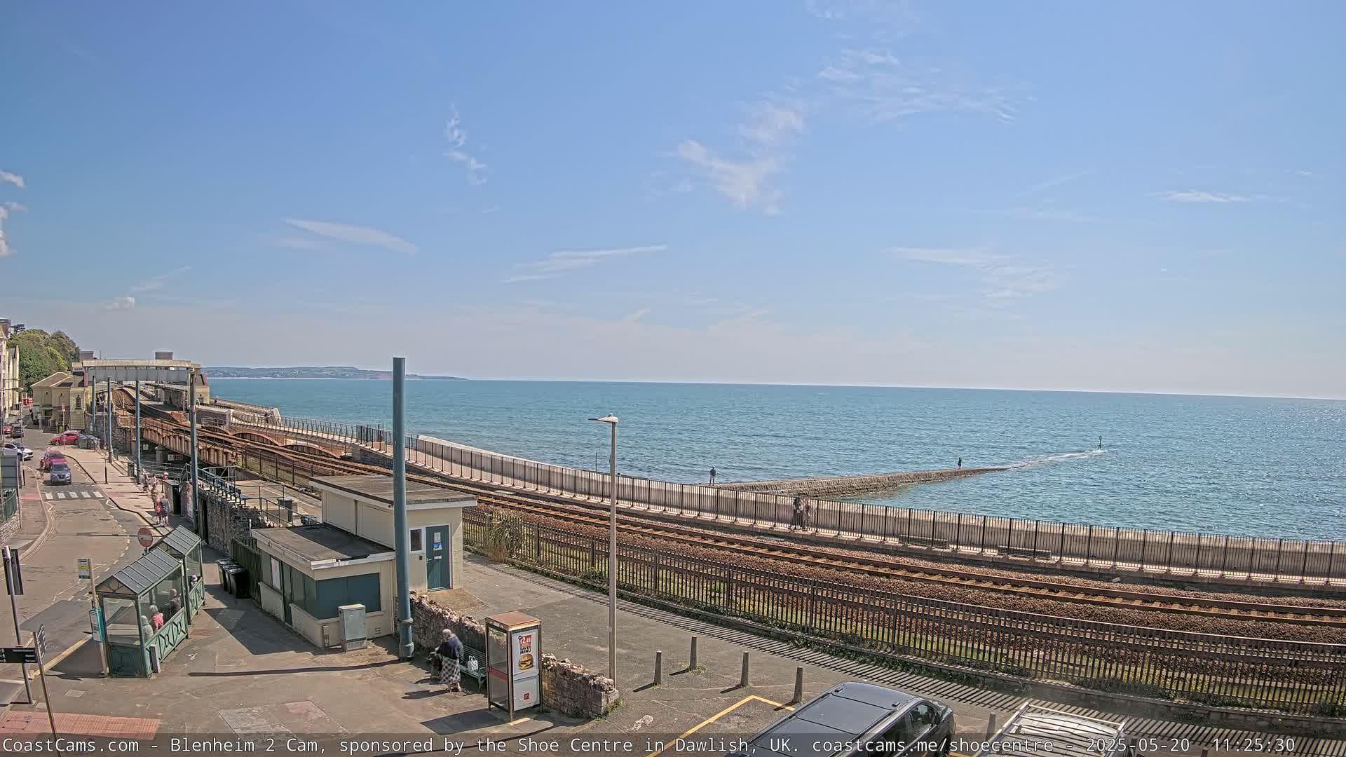 A coastal train track runs alongside a seawall overlooking a calm ocean under a mostly clear blue sky.