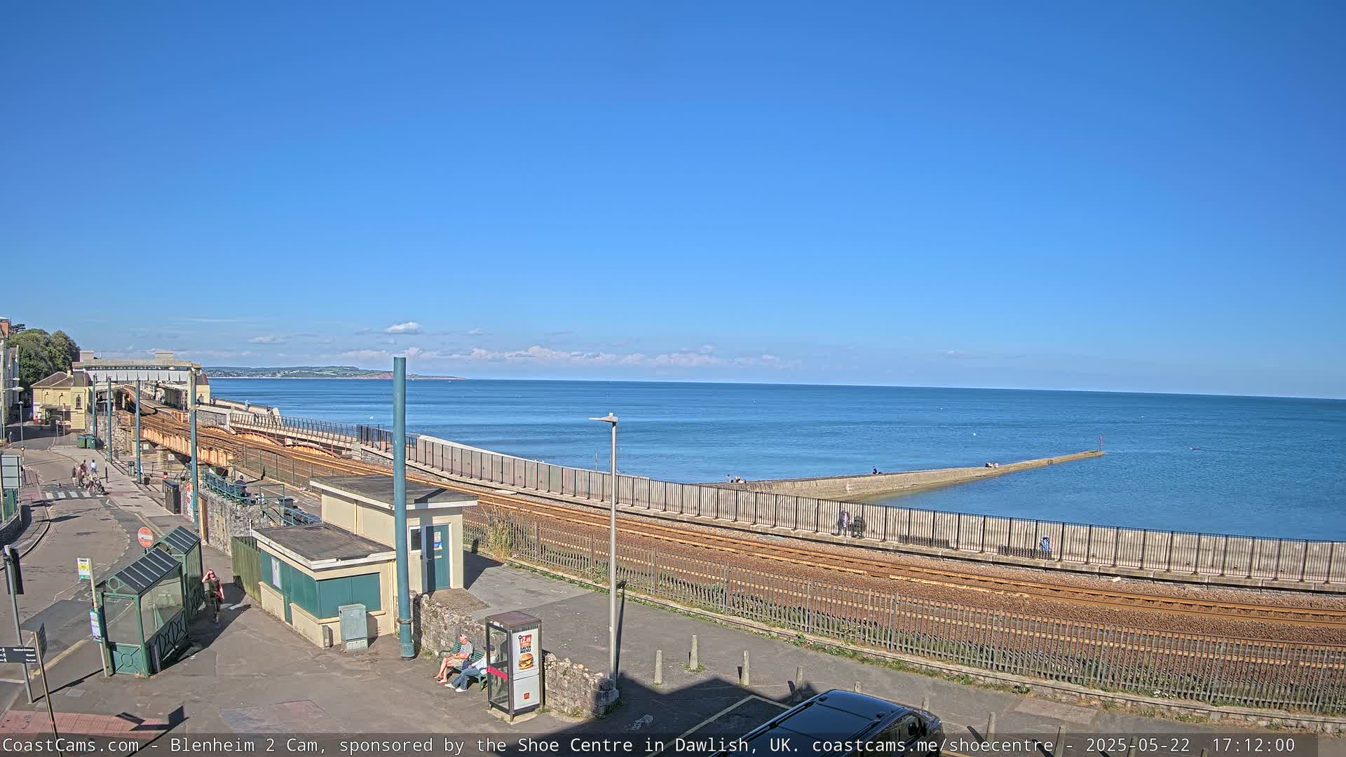 A seaside train track runs alongside a seawall, with a calm blue sea and clear blue sky in the background.