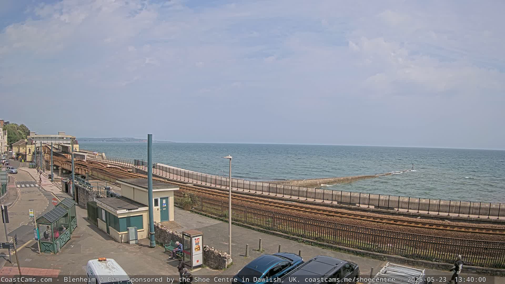 A seaside train track curves along a seawall, overlooking a calm ocean under a partly cloudy sky.