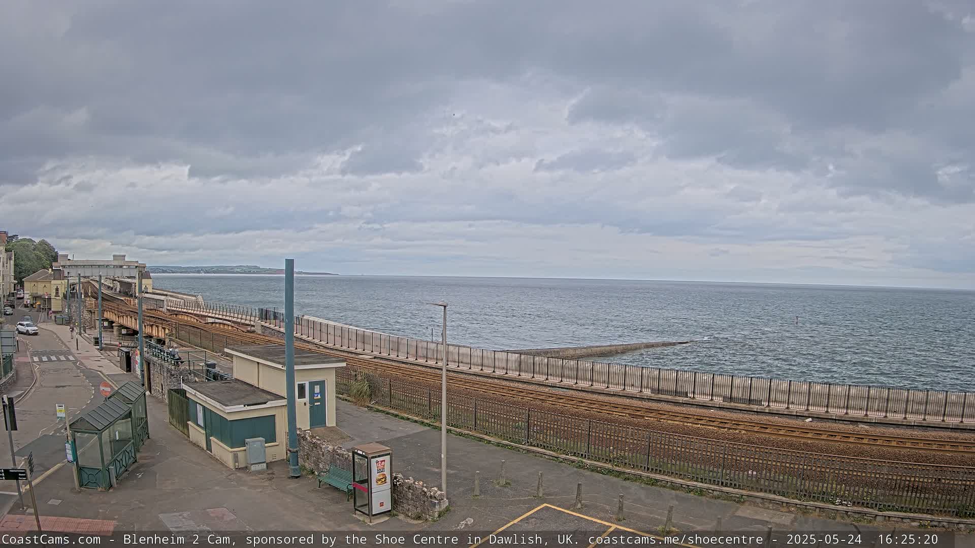 A train track runs alongside a seawall next to a calm, grey sea under an overcast sky.